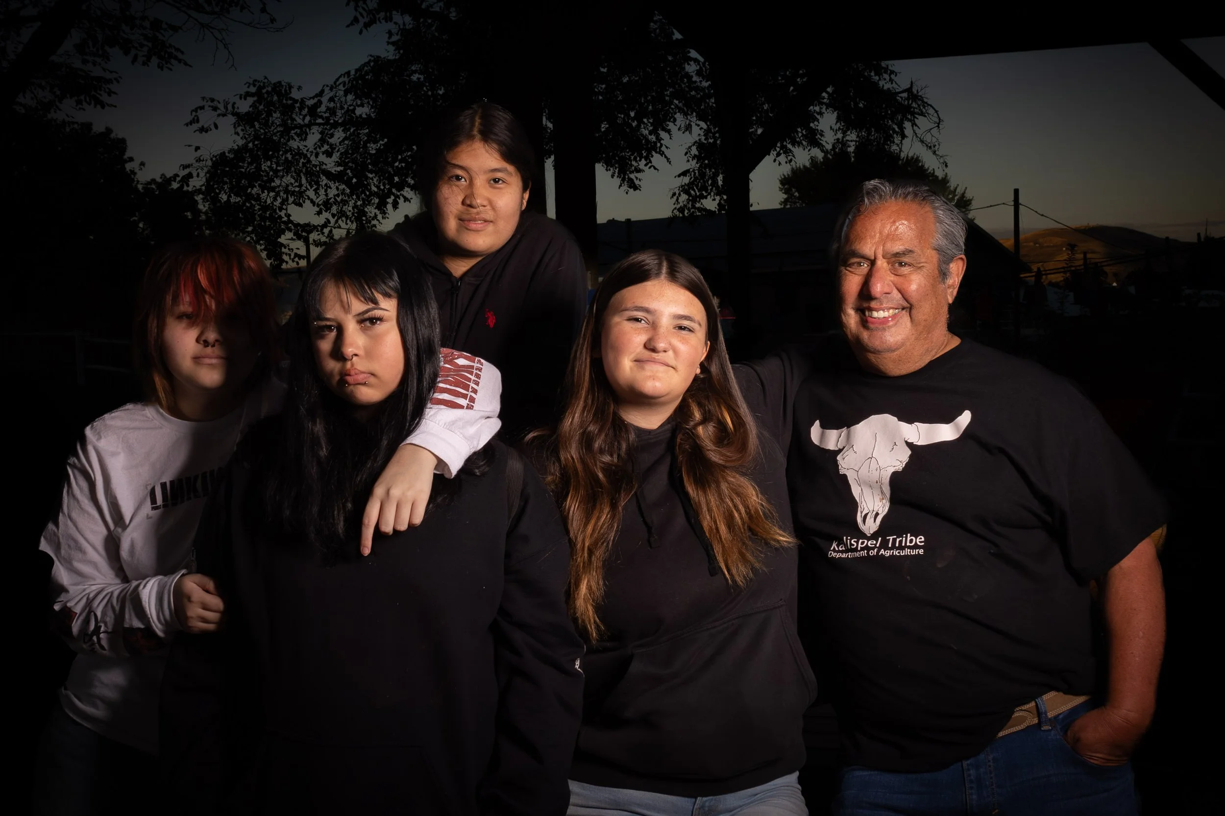 A group of five people standing outdoors during dusk with trees and hills in the background. Two young girls are in the front, one with dark hair and the other with light brown hair, both smiling. Behind them, a young man with black hair and a black hoodie, and a smiling older man with grey hair wearing a black t-shirt with a ranching graphic and the text 'Kalispell Tribe Department of Agriculture.' The group appears happy, posing for a photo.