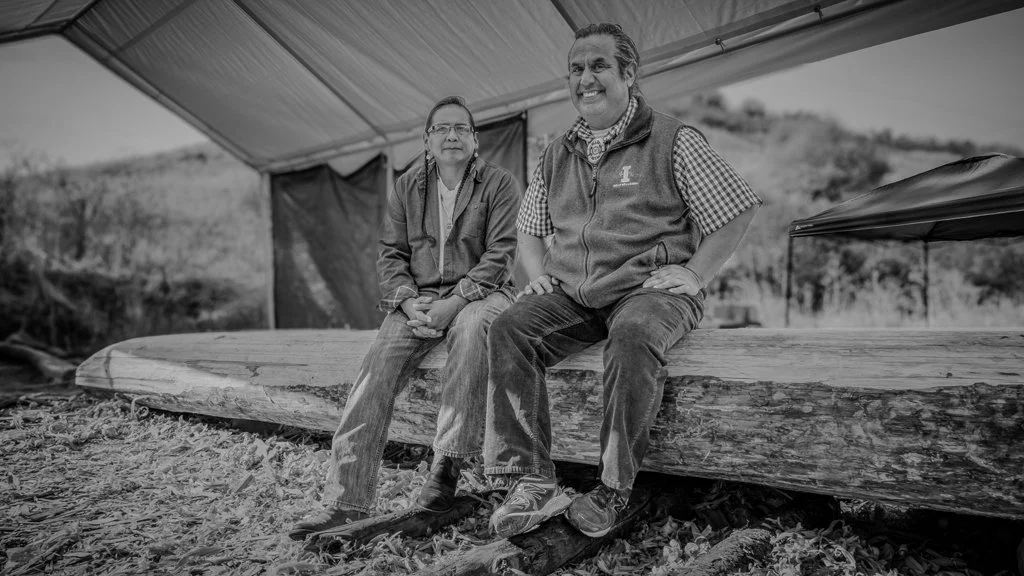 Two people sitting on a large log in front of a tent outdoors in black and white.