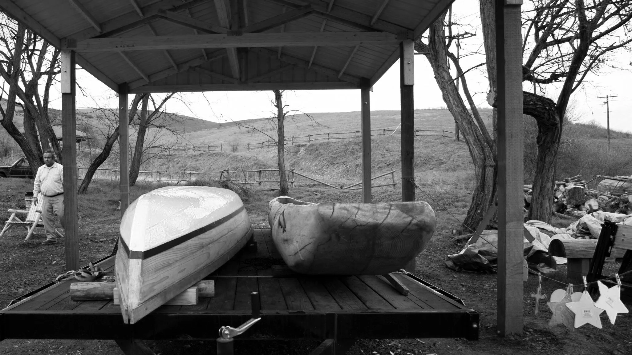 Black and white photo of a park or outdoor area with trees, a wooden pavilion, kayaks, and a man standing near a bicycle.