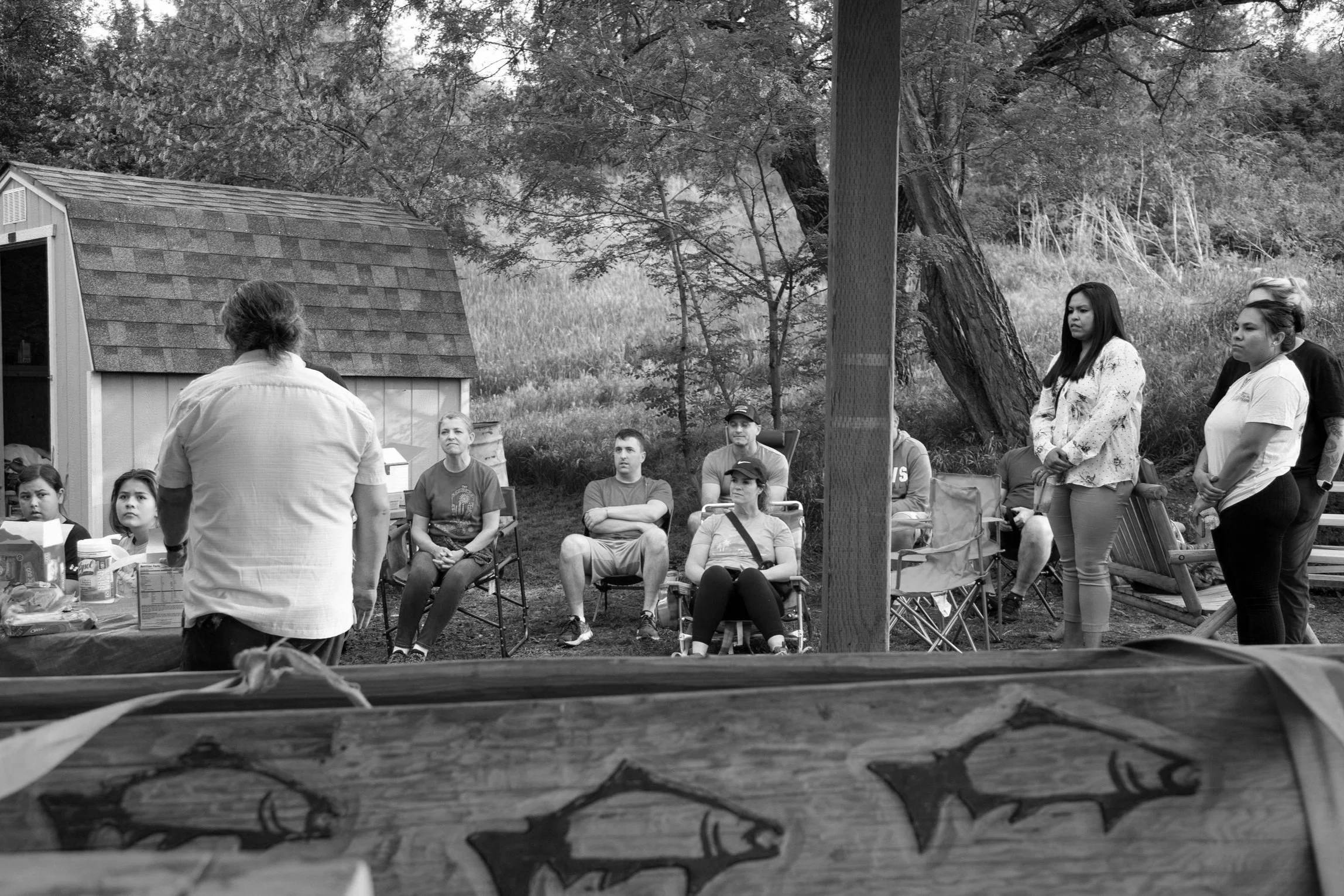 A group of people gathered outdoors for a meeting or presentation. Several individuals are seated on chairs, attentively listening, while others stand nearby, in front of a small building with a shingled roof. The setting is natural with trees and gr