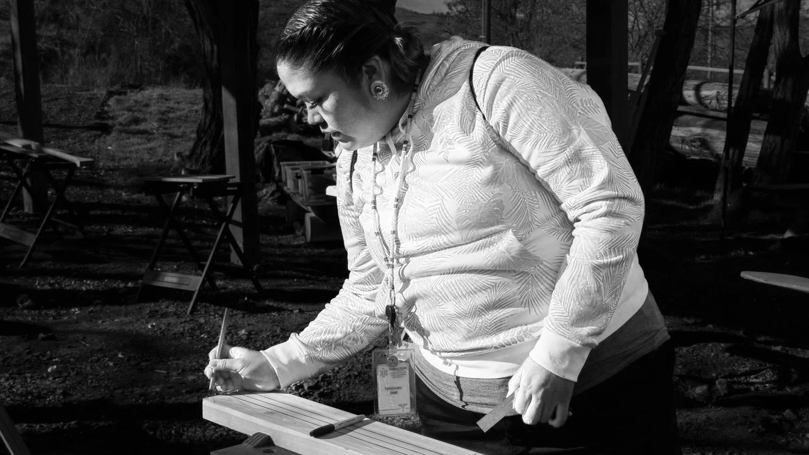 A woman wearing a patterned hoodie and earrings, with an ID badge around her neck, is writing on a piece of wood with a pen outdoors with trees and tables in the background.