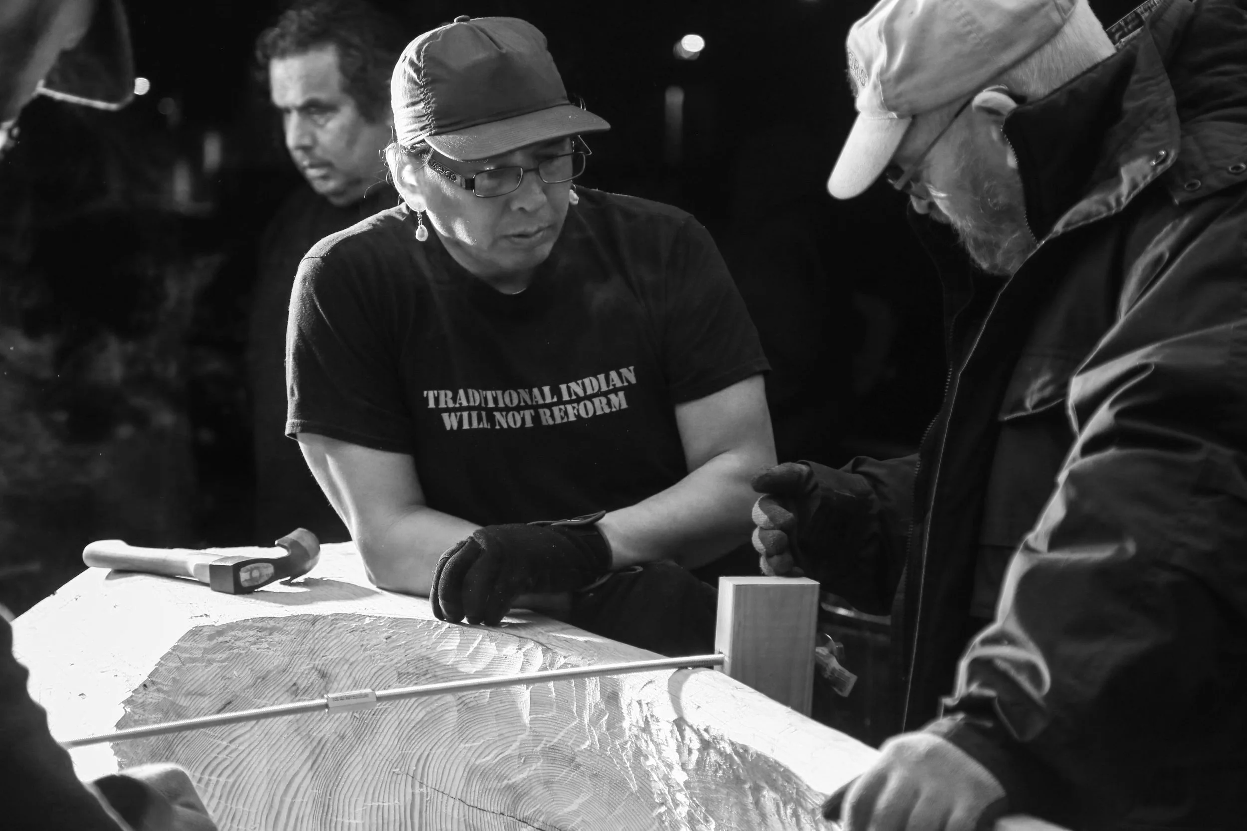 A group of people, including a woman with glasses and a cap, are gathered around a table working on a woodworking project. The woman is wearing a shirt that says 'TRADITIONAL INDIAN WILL NOT REFORM' and is focused on her task.