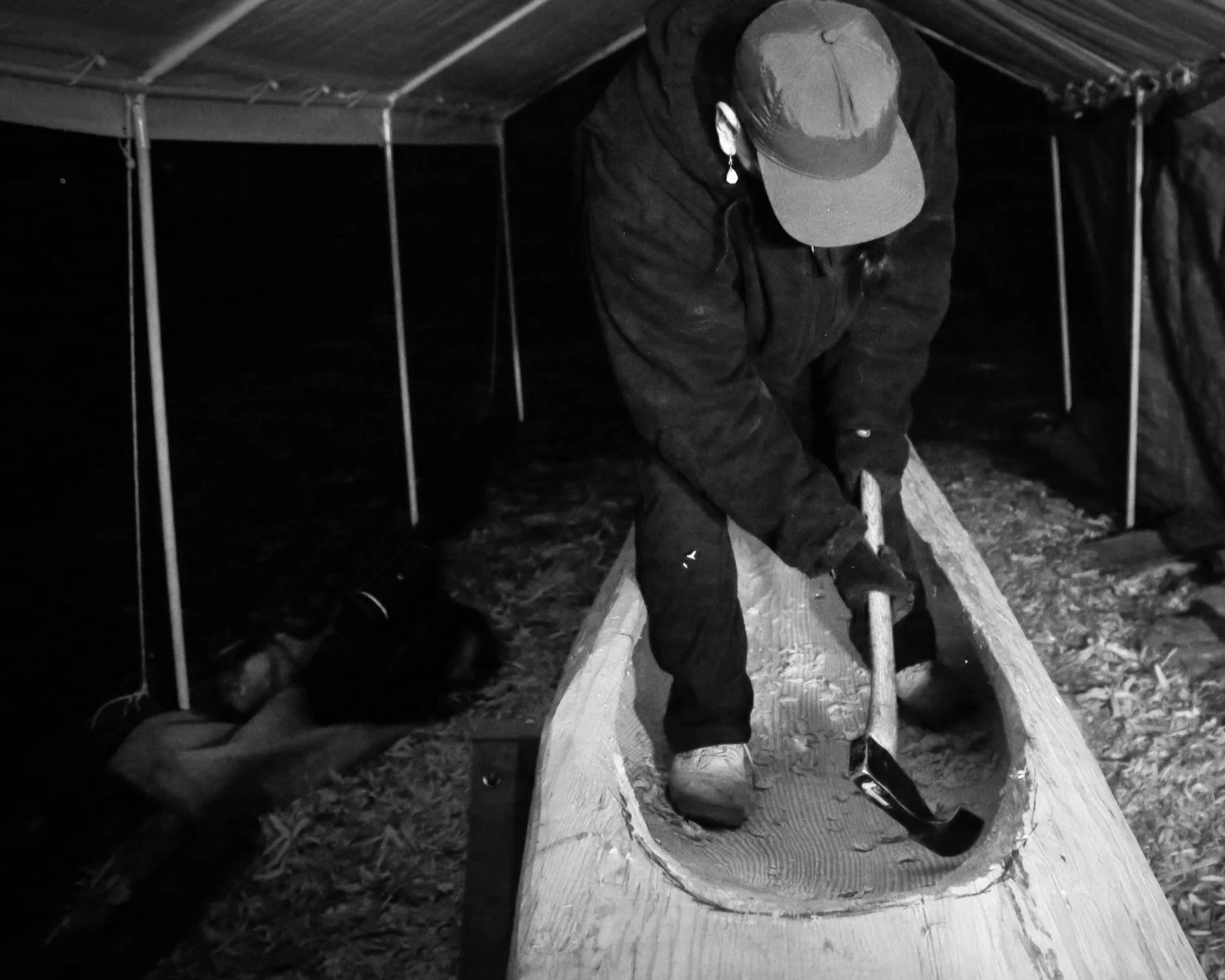 A person wearing a hat and earrings is working on a wooden project outdoors at night, under a canopy. They are bending over a large piece of wood on a wooden sawhorse, using a hand tool, possibly a saw.