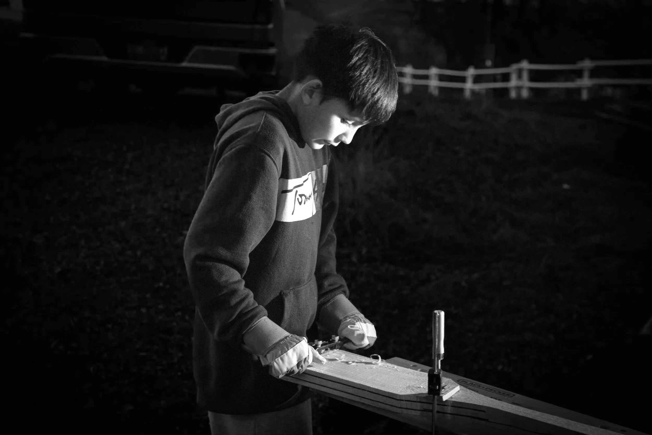 A young boy wearing a hoodie and gloves is working on a woodworking project outdoors at night, using a handsaw on a piece of wood secured on a worktable.