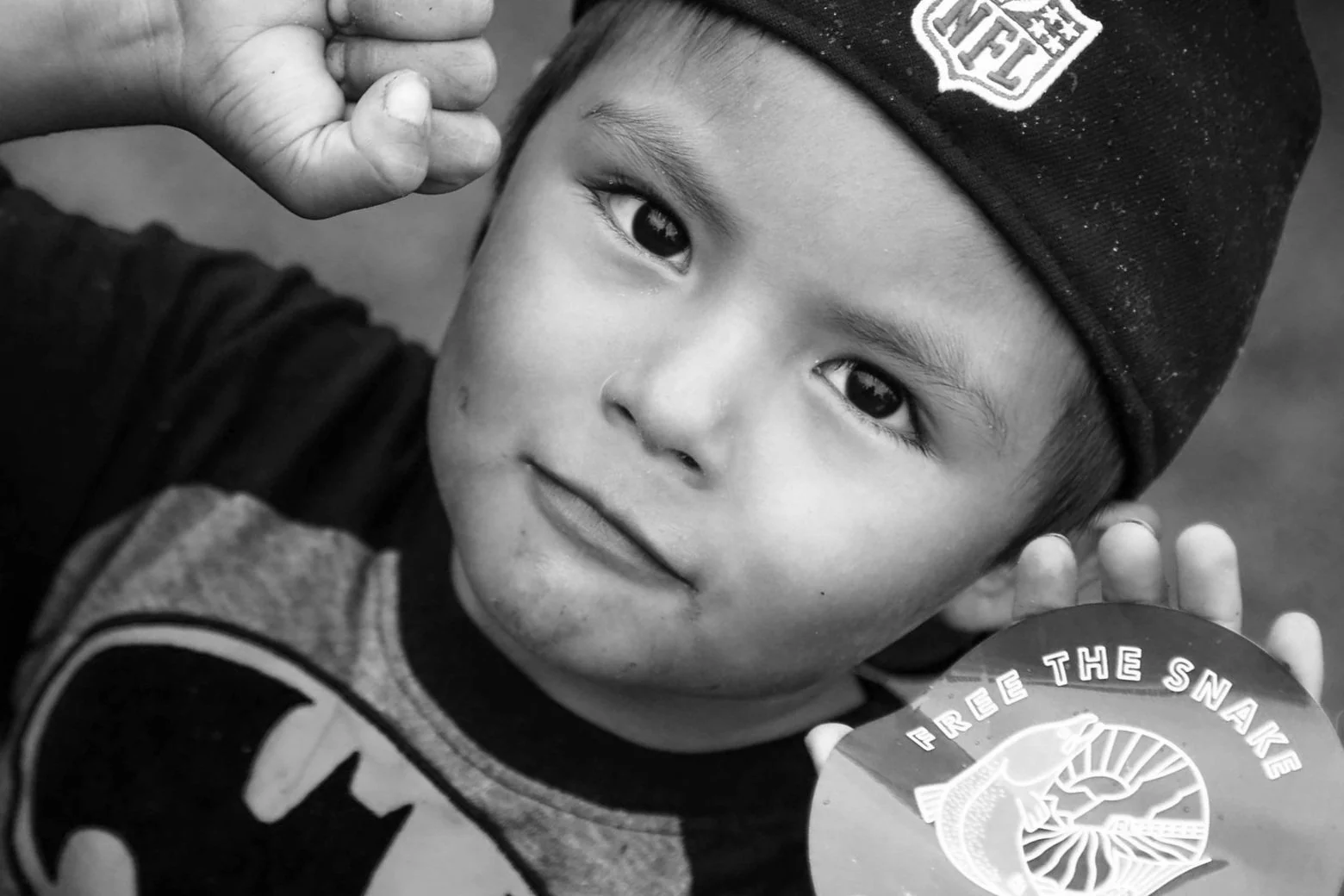 Close-up black and white photo of a young boy wearing a beanie hat and a t-shirt, holding a circular medal with the words "FREE THE SNARES" and an image of a snake.