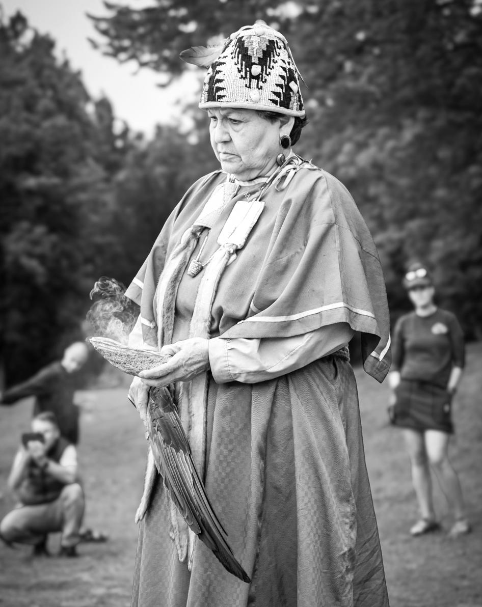 A woman dressed in traditional Native American clothing stands outdoors, holding a burning object. She wears a patterned hat and jewelry, with trees in the background. Two children and a woman are seen in the blurred background.