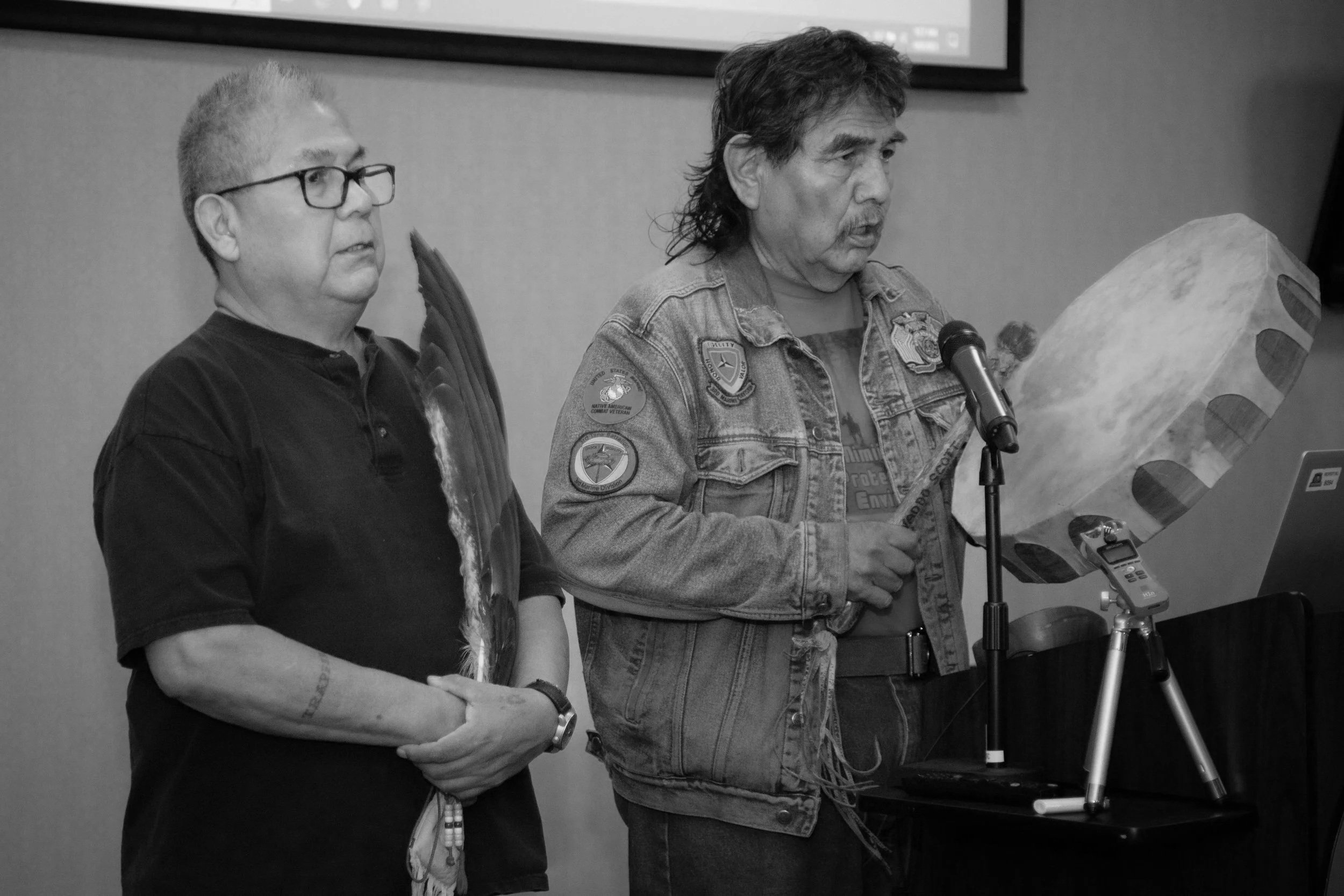 Two men standing at a table with a microphone. One is reading from a large piece of paper, the other looking on. Both are wearing casual clothing.