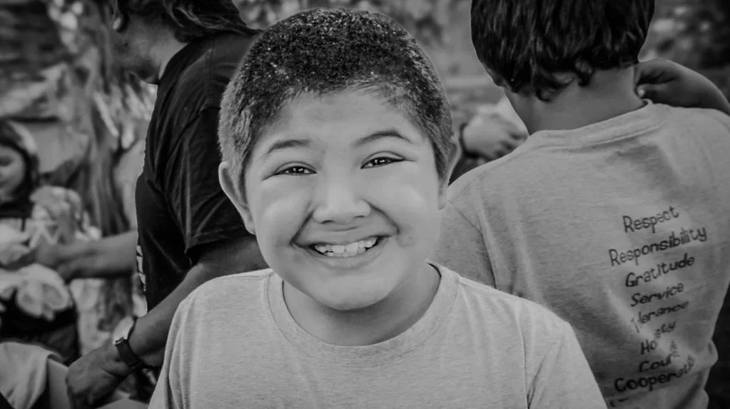 A smiling young boy with short hair in the foreground, surrounded by other children, one of whom is wearing a T-shirt with words like Respect, Responsibility, Gratitude, Service, Love, and Co-operation printed on the back, in an outdoor setting.