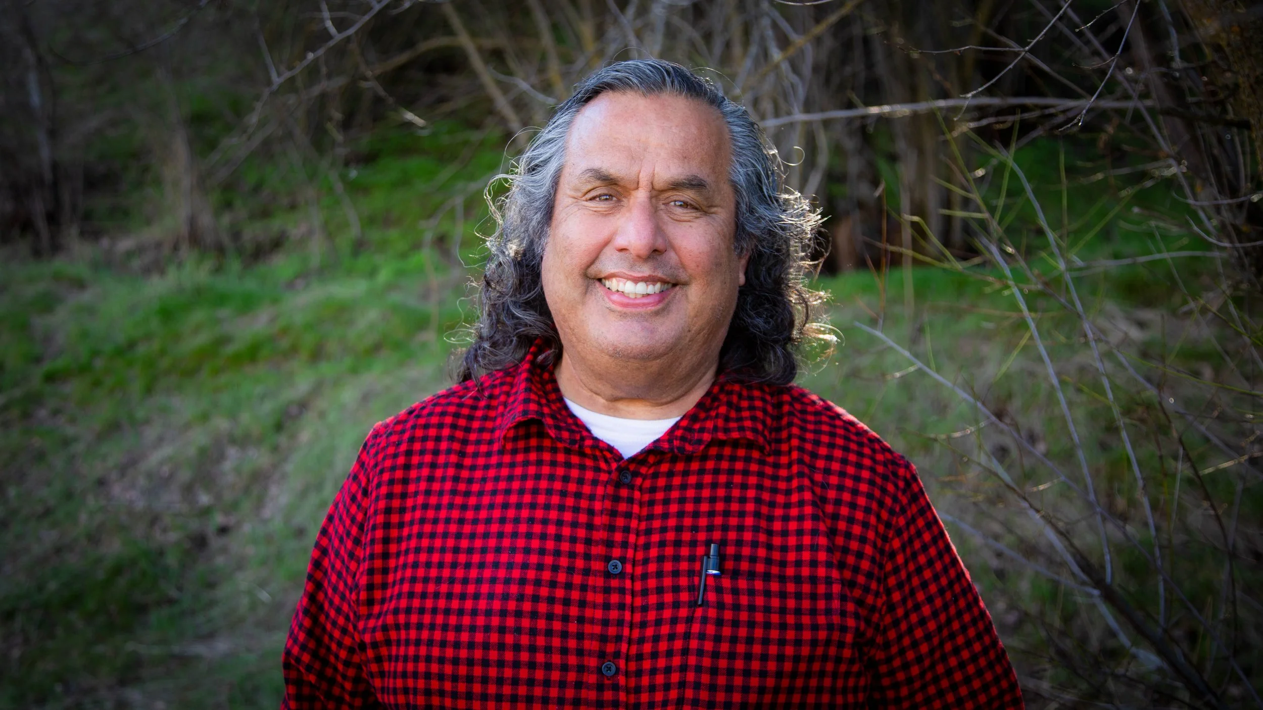 A smiling man with gray hair and a beard, wearing a red and black checkered shirt, standing outdoors with green grass and bushes in the background.