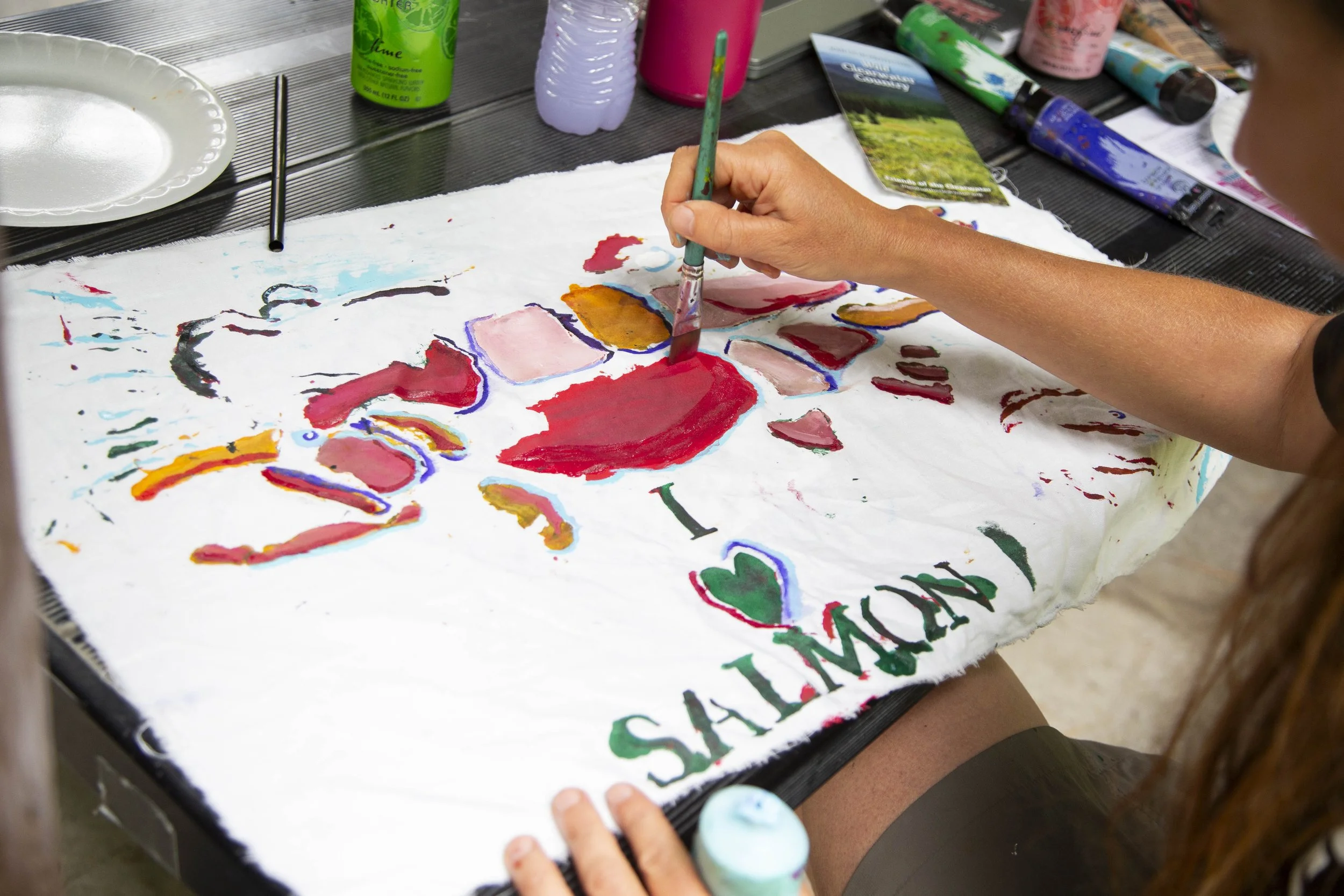 Child painting on a large white cloth with colorful paints, surrounded by art supplies on a table.