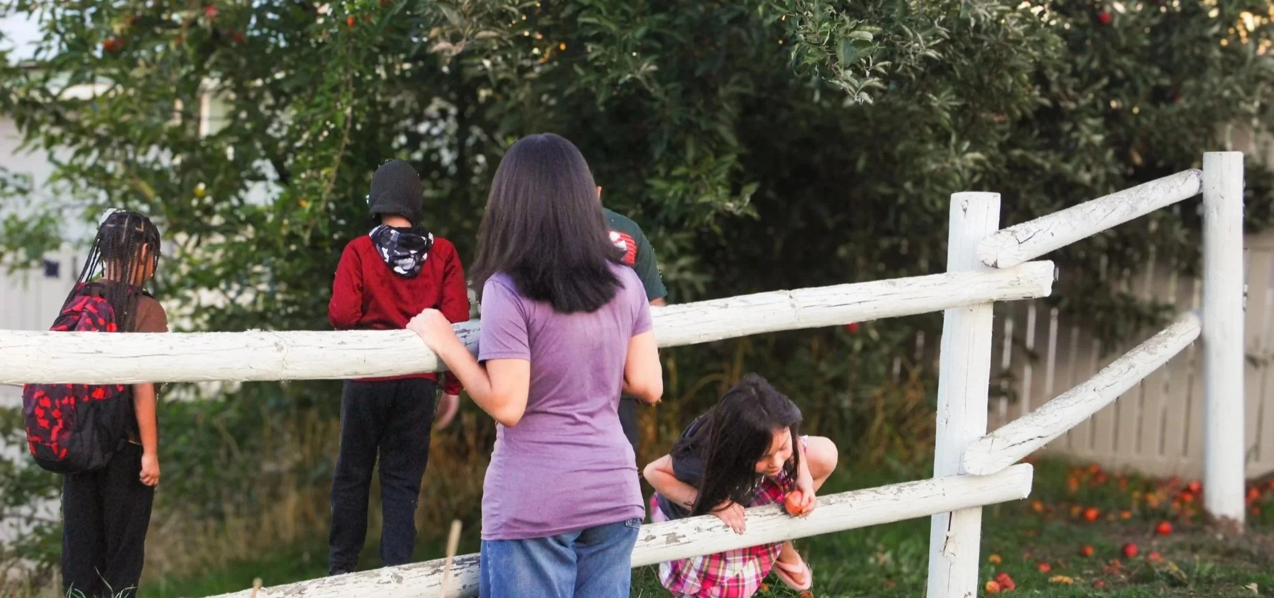 A woman and four children near a white wooden fence with greenery and trees in the background.