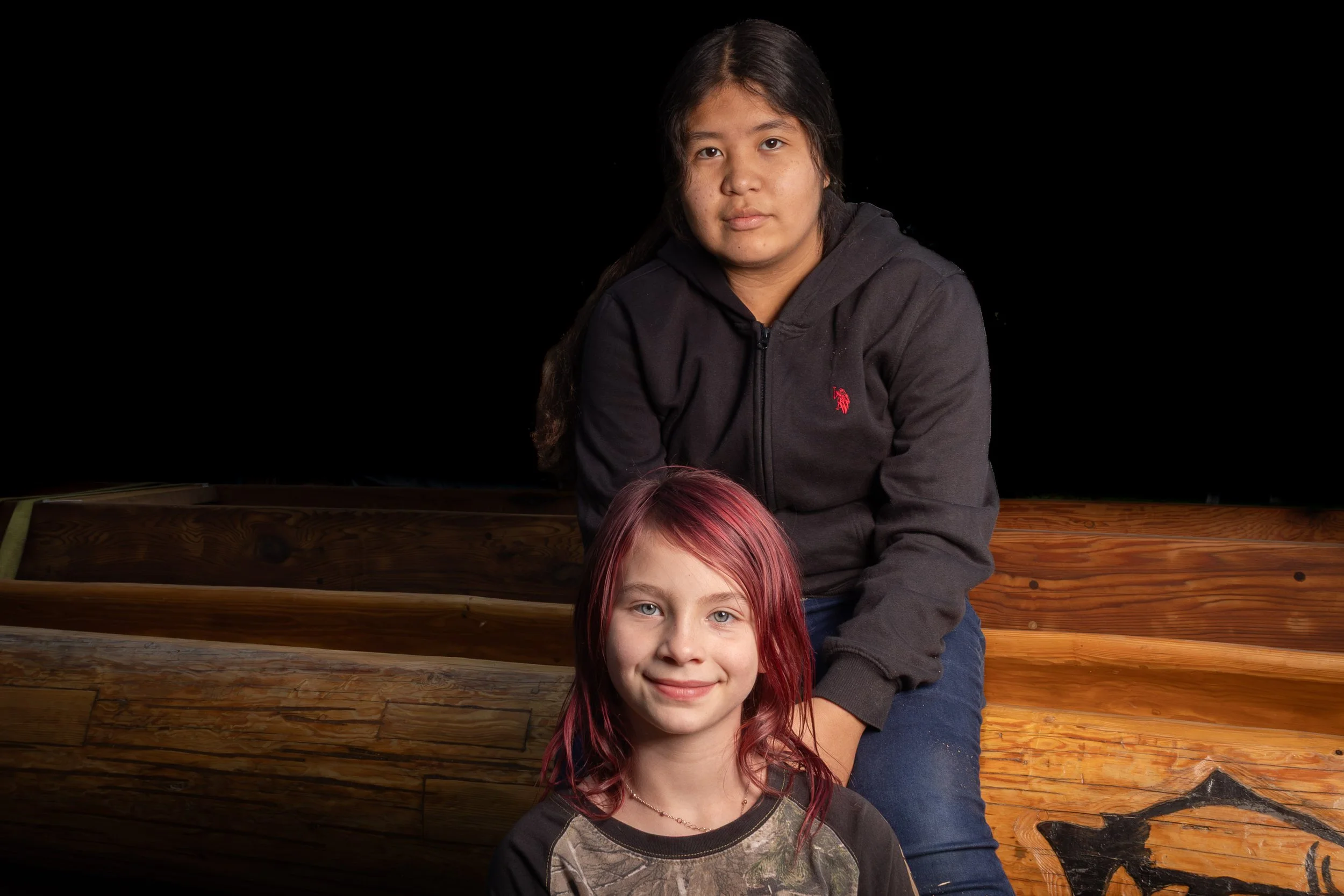 Two girls, one with dark hair and one with red hair, smiling at the camera against a black background. They are sitting on wooden steps.