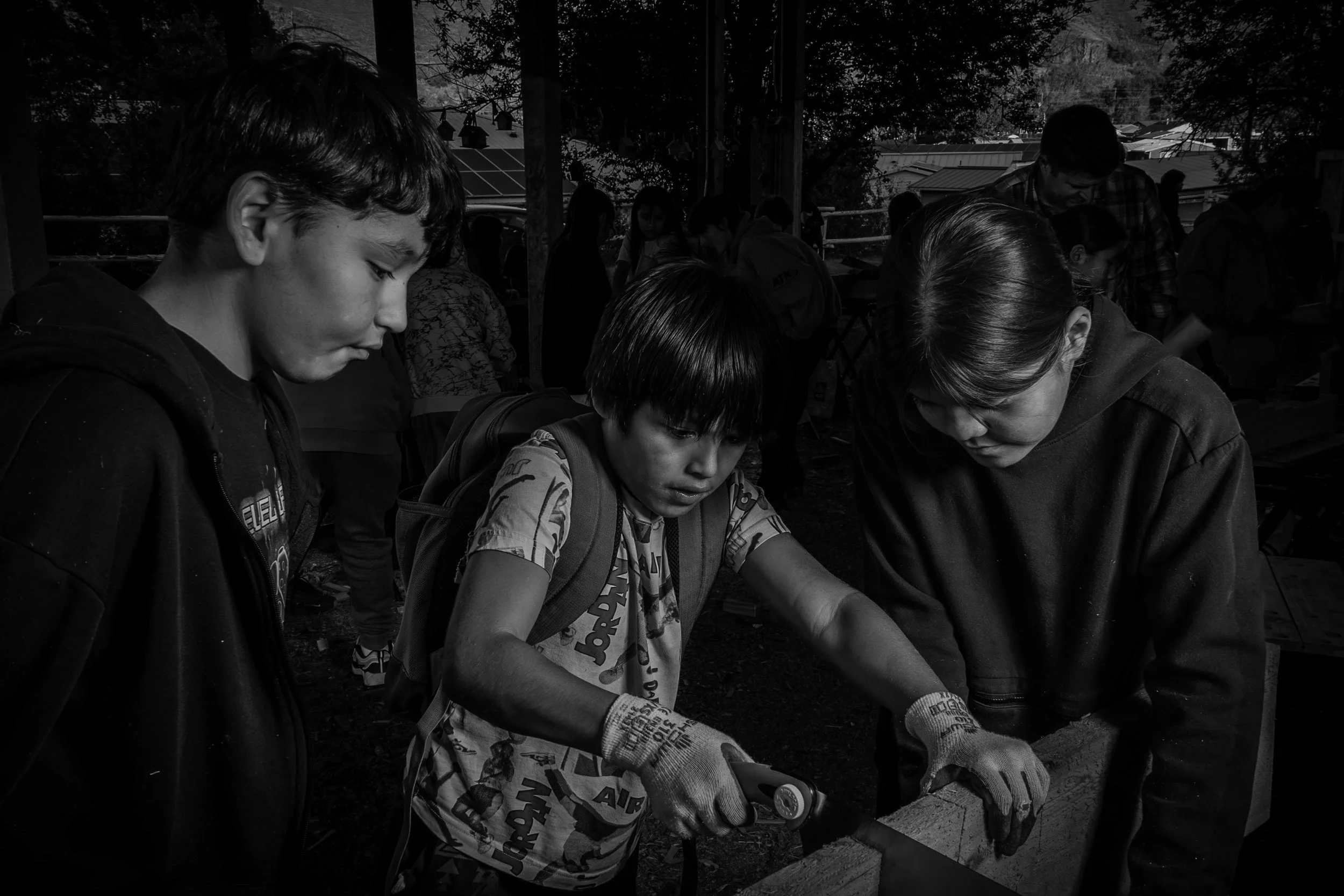Group of children and adults working together at an outdoor table, focused on a project, under a shaded structure with trees and rooftops in the background.
