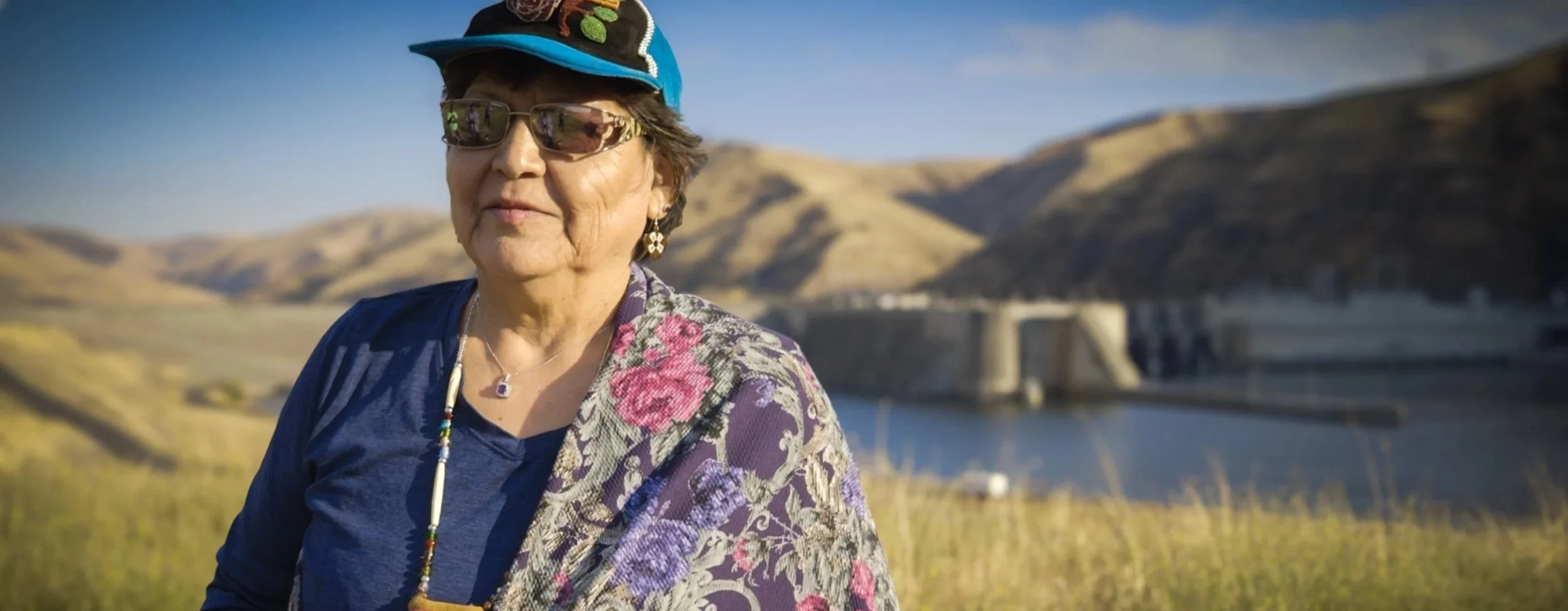 An older woman wearing sunglasses, a hat, and floral-patterned clothing standing outdoors near a body of water and hills, with a dam structure in the background.