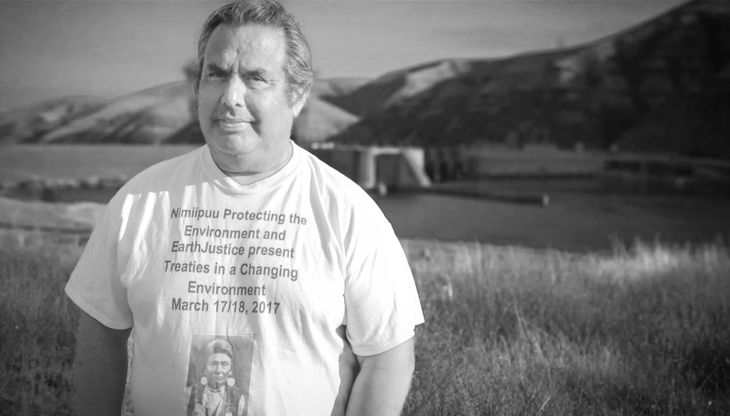 A man standing outdoors with rolling hillside background. He is wearing a white t-shirt with text related to environmental protection and a photo of a person. The image is in black and white.