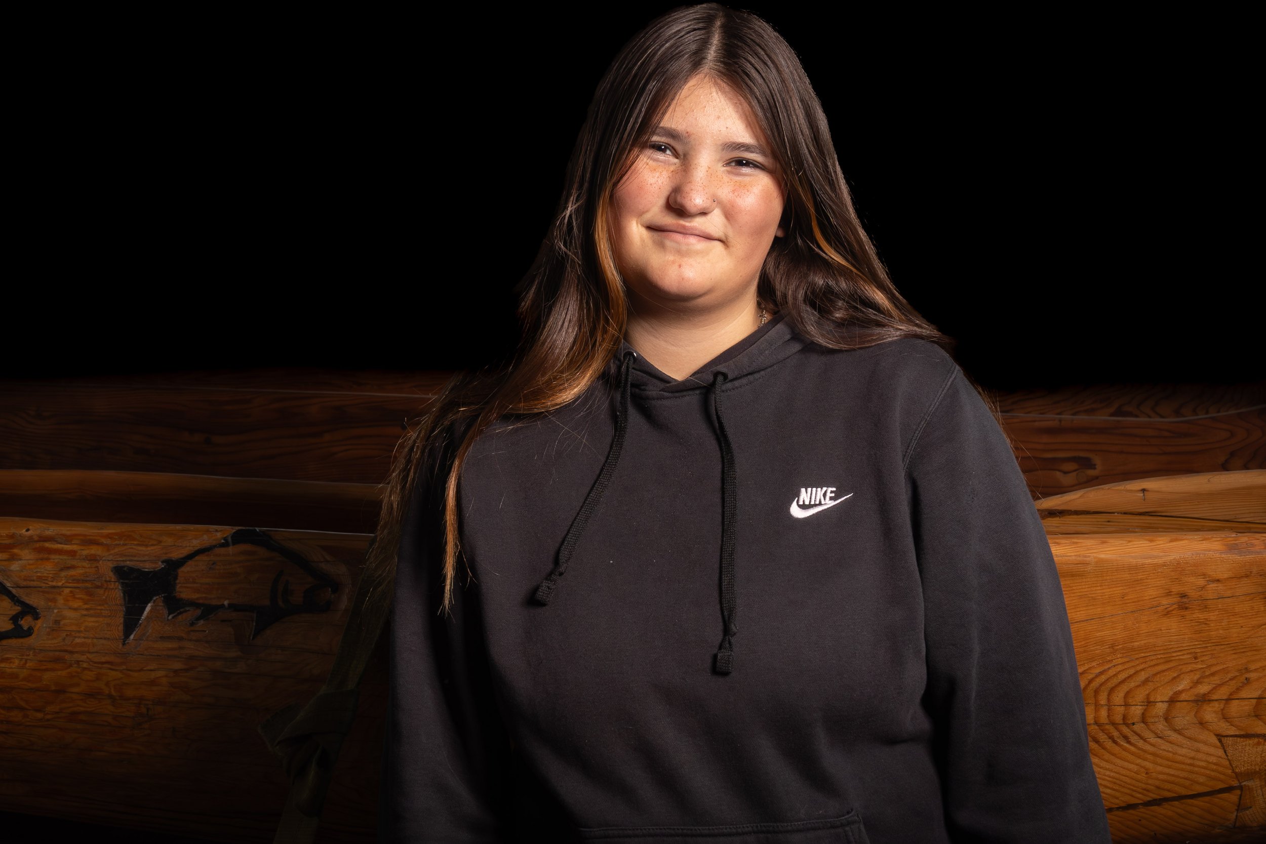 A young woman with long brown hair, freckles, and a nose piercing wearing a black Nike hoodie standing in front of a wooden background with graffiti.