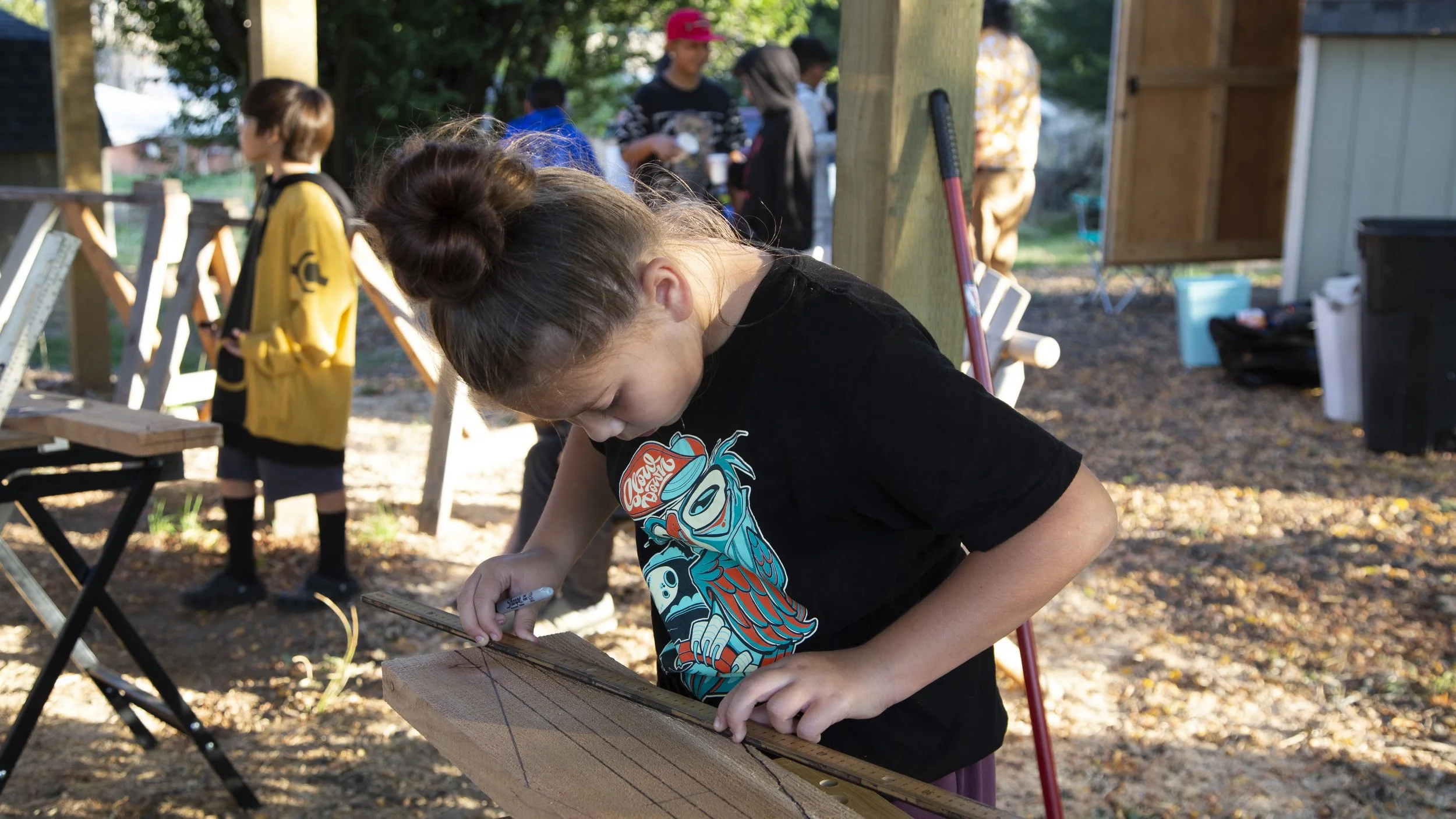 Young girl with a bun hairstyle wearing a black T-shirt with a colorful graphic, working on a woodworking project outdoors, surrounded by other children and adults, with tools and supplies visible in the background.