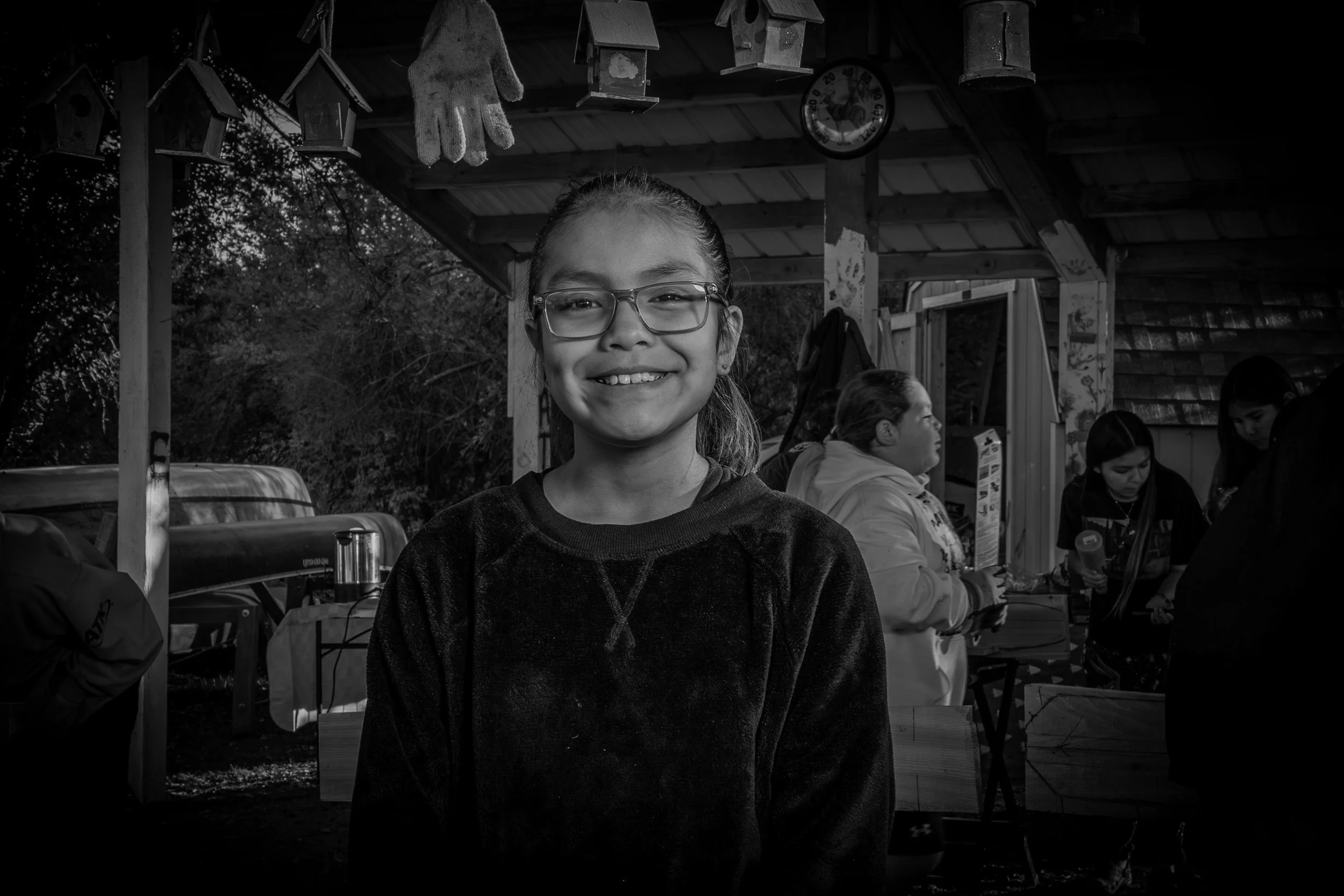 A girl with glasses smiling at the camera outdoors at a gathering with people and birdhouses hanging above, in black and white.