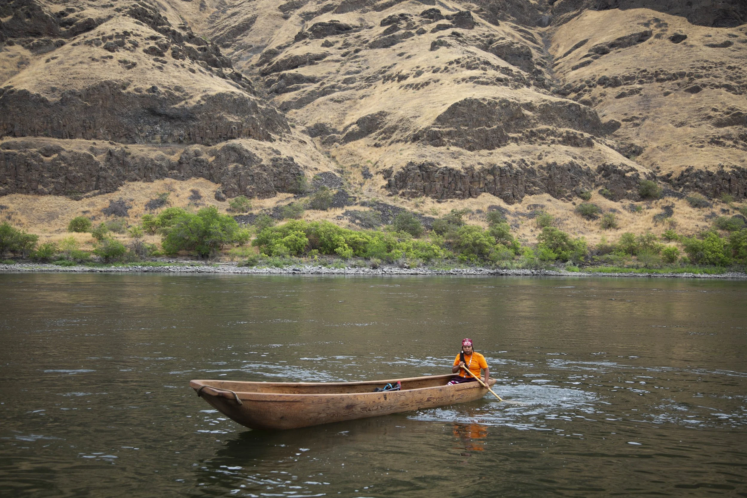 A person in an orange shirt and pink headscarf sits in a wooden boat on a river, with steep, dry brown hills in the background and green shrubs near the riverbank.