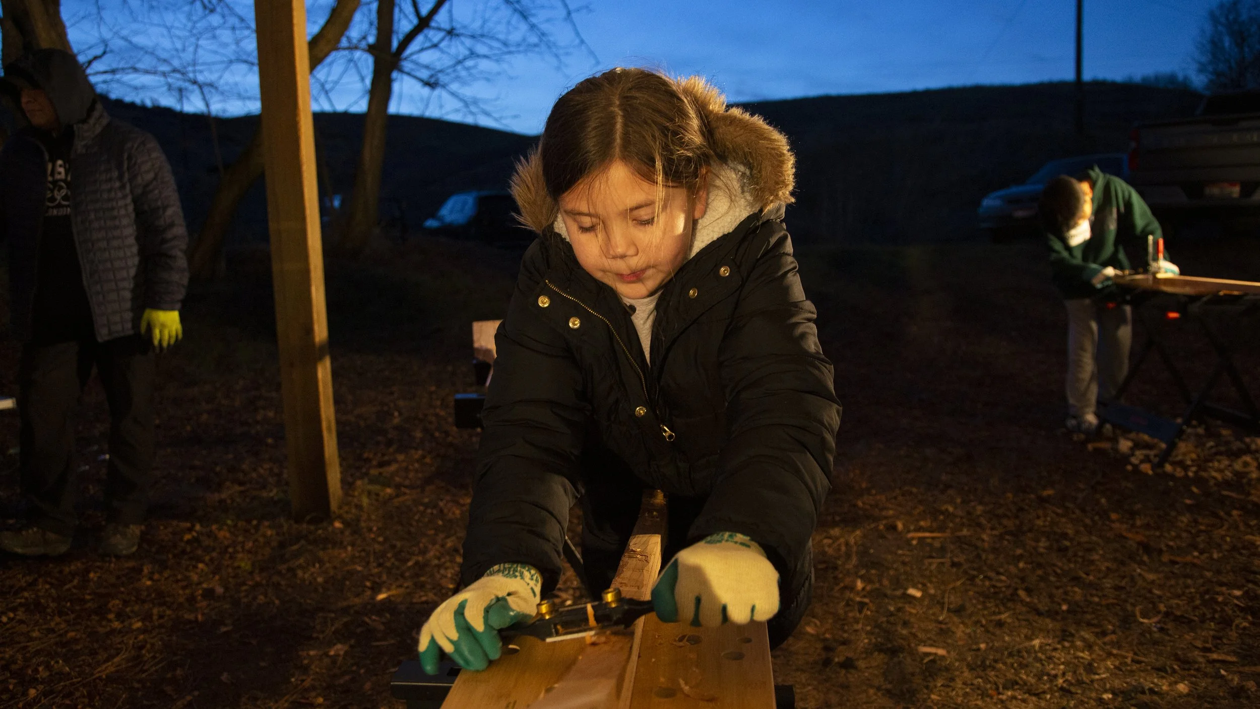 A young girl wearing gloves and a black jacket is sawing a piece of wood outdoors during dusk. Two other people are in the background, one on the left wearing a hoodie and gloves, and another on the right at a table with tools, all under a wooden she