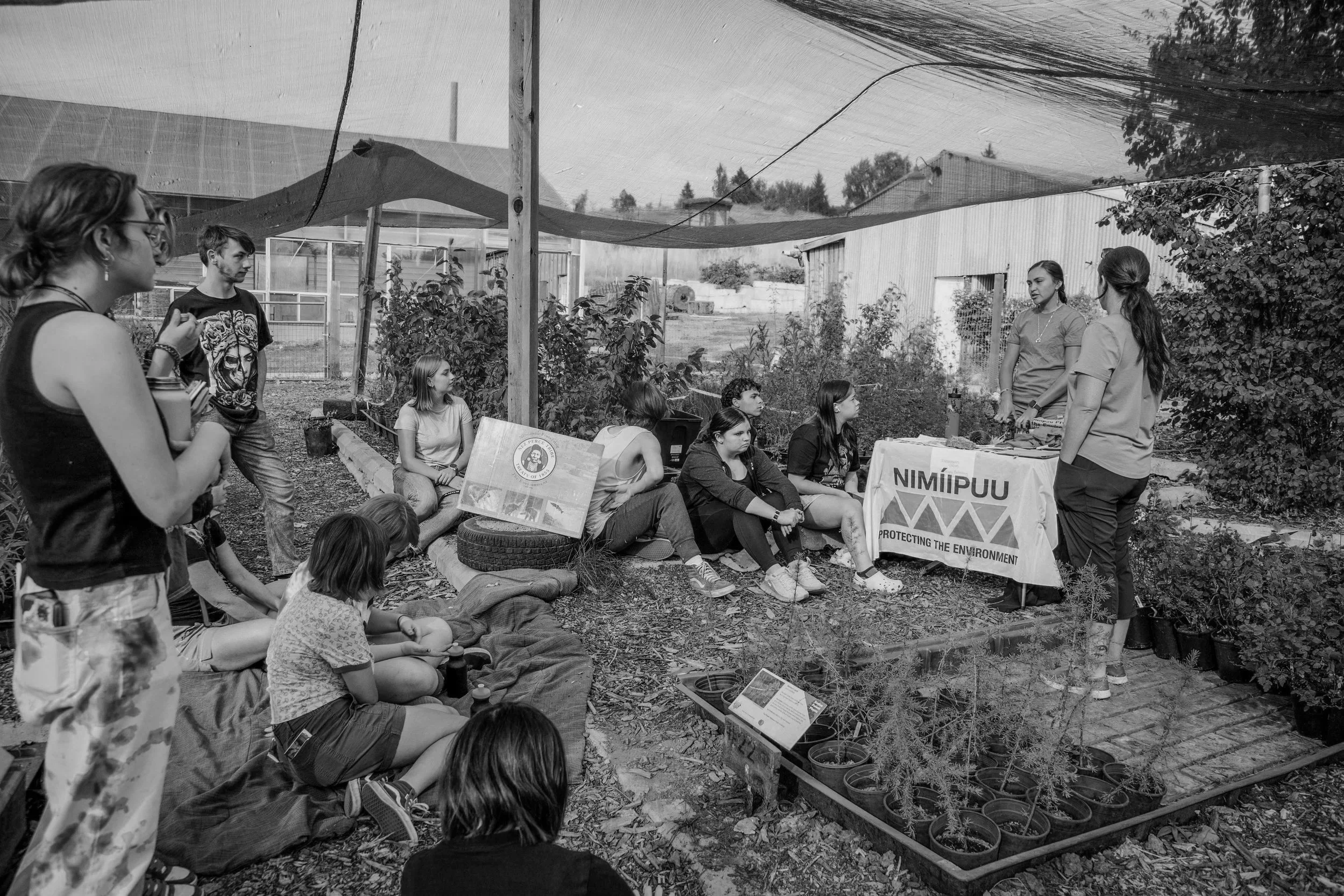 A group of young people gathered outdoors in a garden, listening to a woman speaking at a table with a banner that reads 'NIMIPUU' and 'Protecting the Environment.' The area is shaded with a canopy, and there are plants and informational signs present.