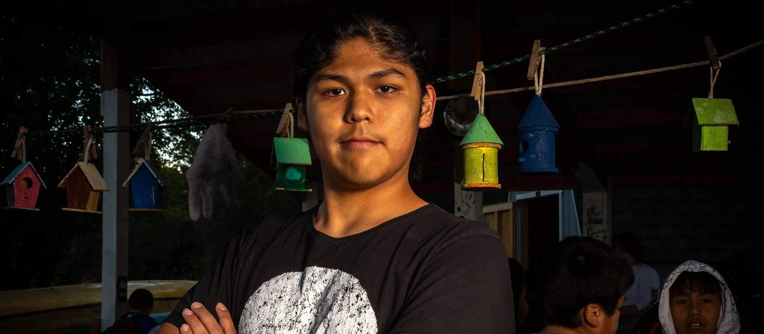 A young man with dark hair and tan skin stands with arms crossed, looking at the camera. Behind him, colorful birdhouses hang from a line under a covered outdoor area, with trees and a person in the background.