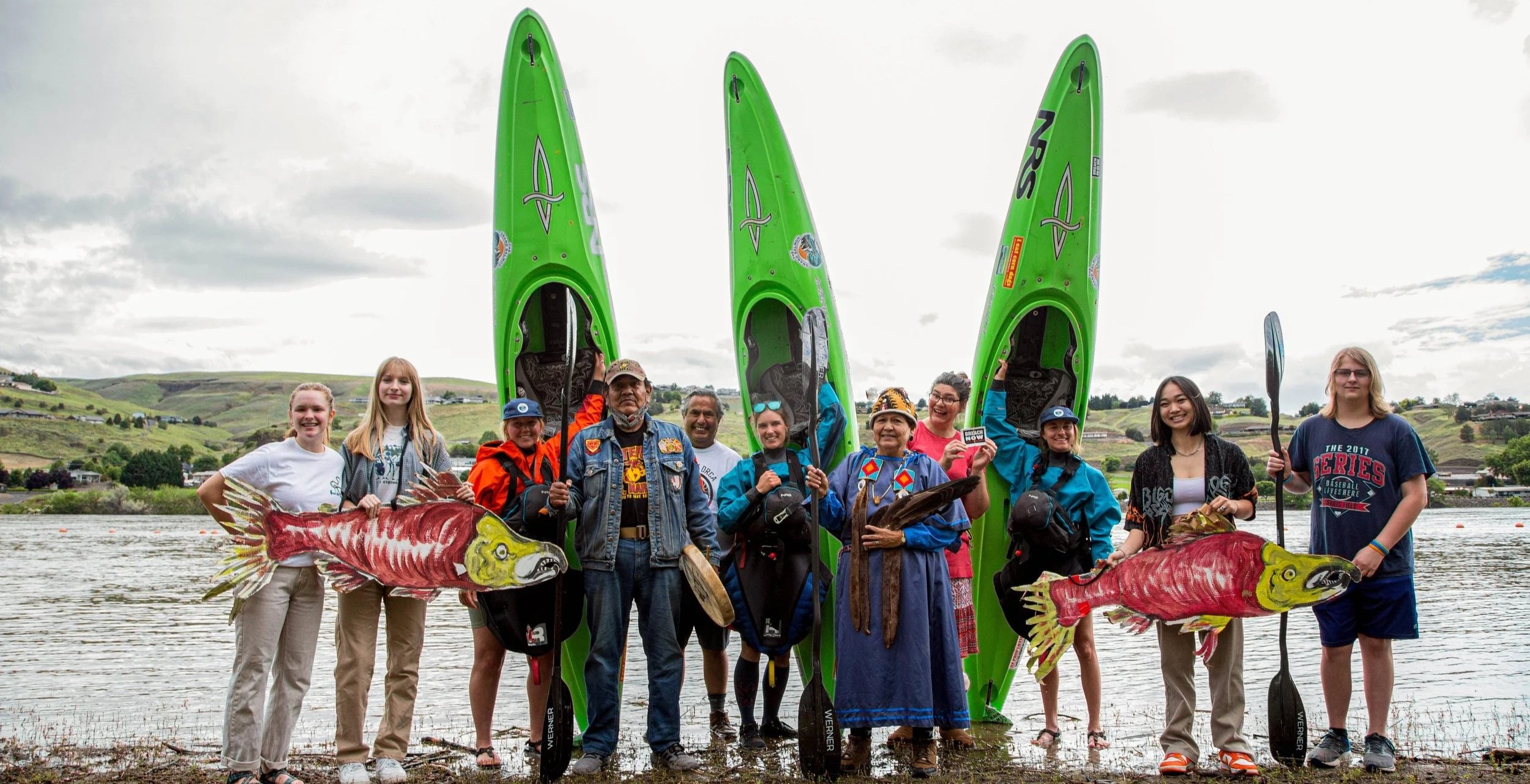 Group of people standing by the water holding kayaks and paddles, with two large green kayaks standing vertically behind them, hills and cloudy sky in the background.