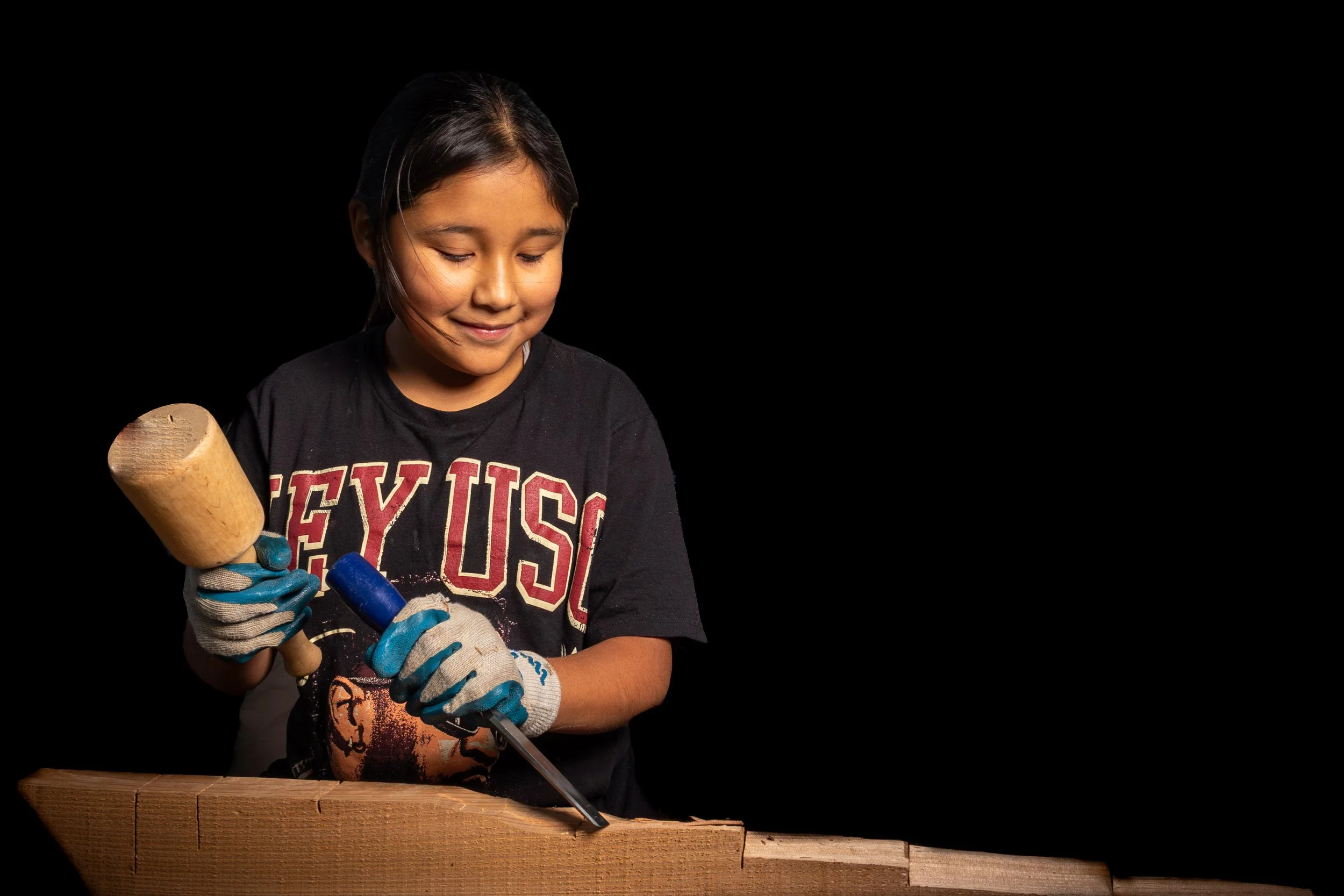 A young girl wearing a black T-shirt with the word 'EYES' and a graphic of a lion on it, holding a sledgehammer and a crowbar, standing behind a wooden surface, against a black background, smiling gently with eyes looking down.