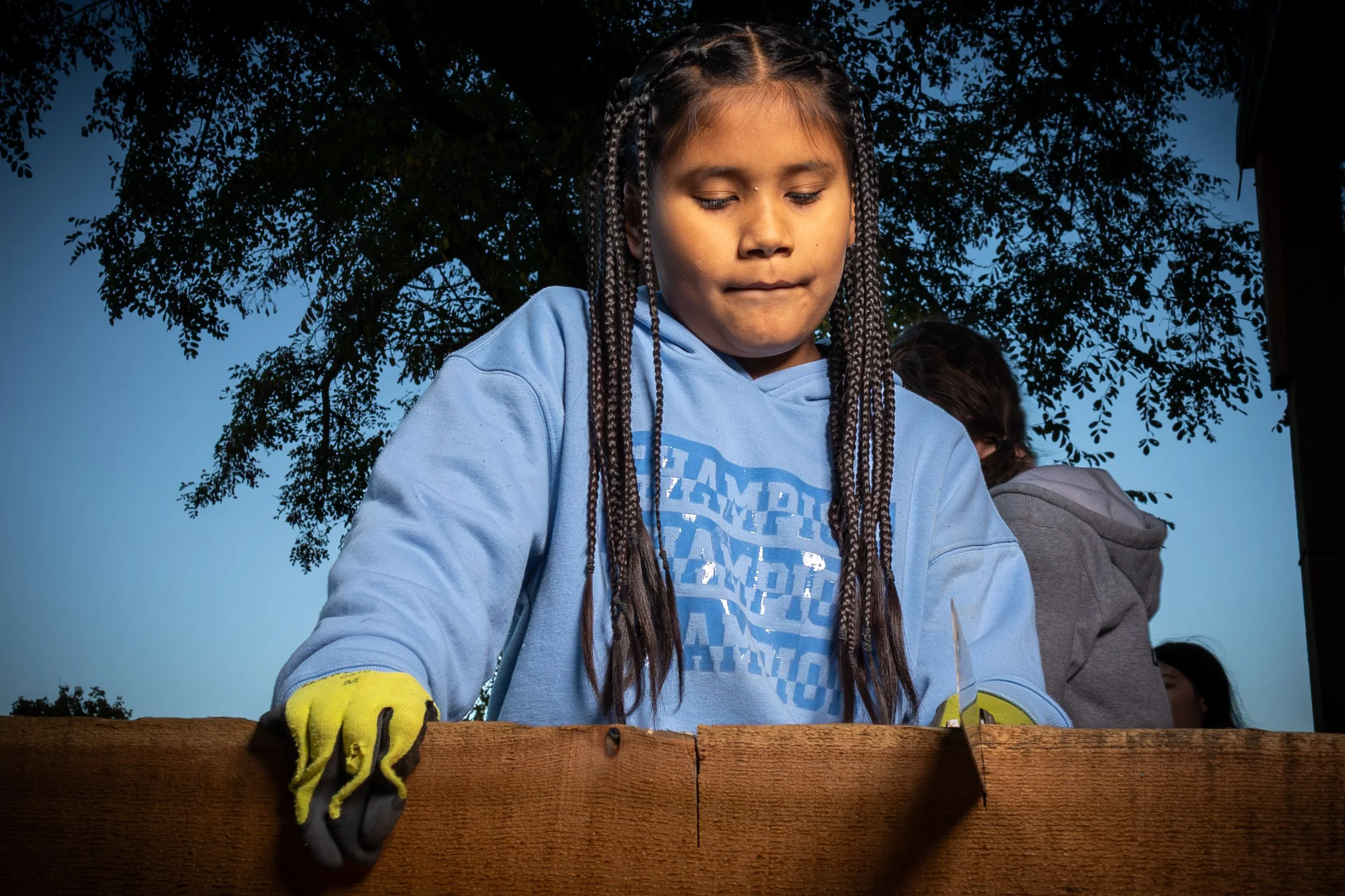 A young girl with braided hair wearing a blue hoodie and yellow gloves is working on a wooden project outdoors during daytime, with trees in the background.