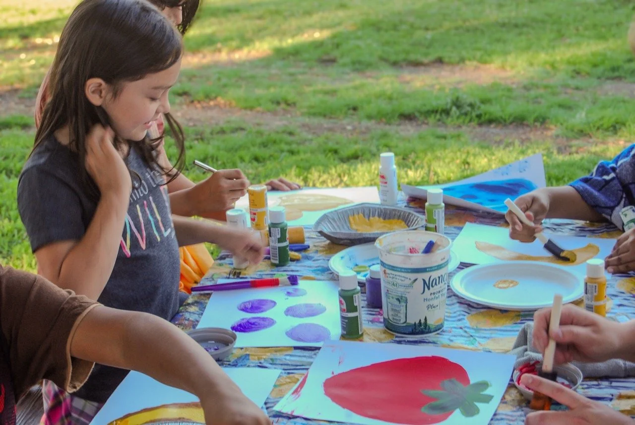Children and adults painting and drawing at an outdoor table on a sunny day.