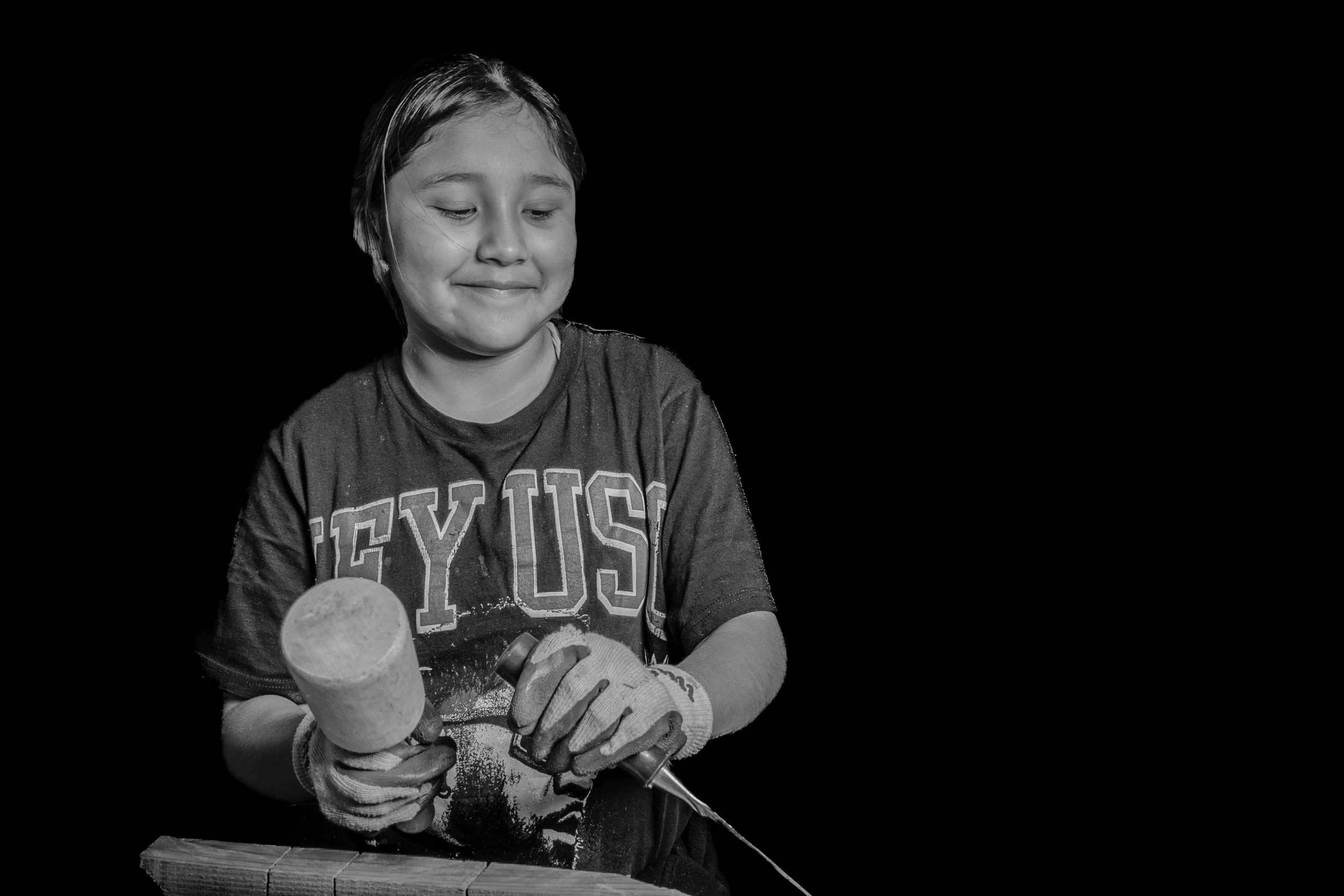 A young girl wearing a Tennessee T-shirt and gloves is holding a wooden bat in her hands, smiling and looking down, with a skateboard on the table in front of her.