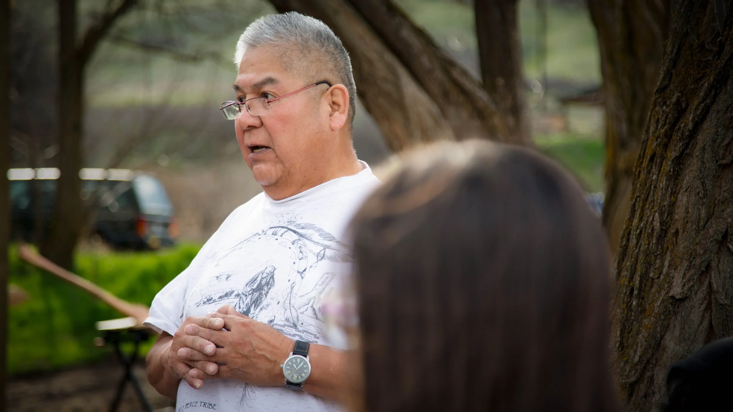 A man with gray hair and glasses, wearing a white t-shirt with a graphic design, stands outdoors with his hands clasped, near a tree. There are blurred cars and greenery in the background.
