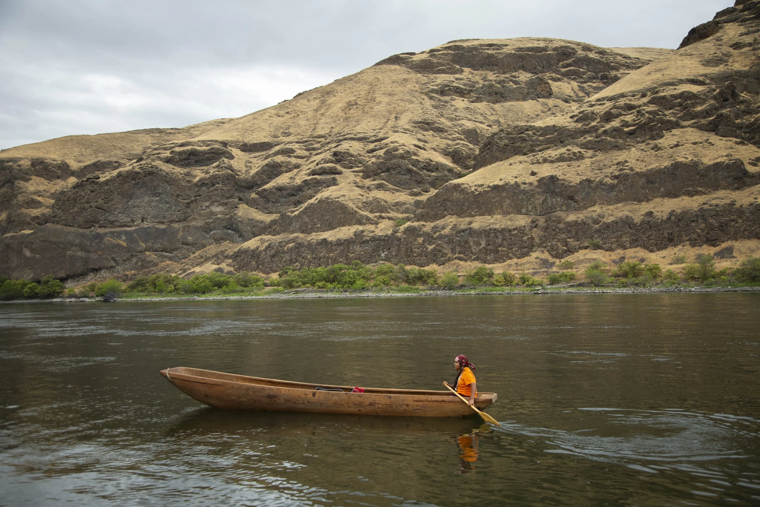 A person in an orange shirt and red headscarf paddles a wooden boat on a calm river, with a large brown and dark green rocky hill in the background and an overcast sky.