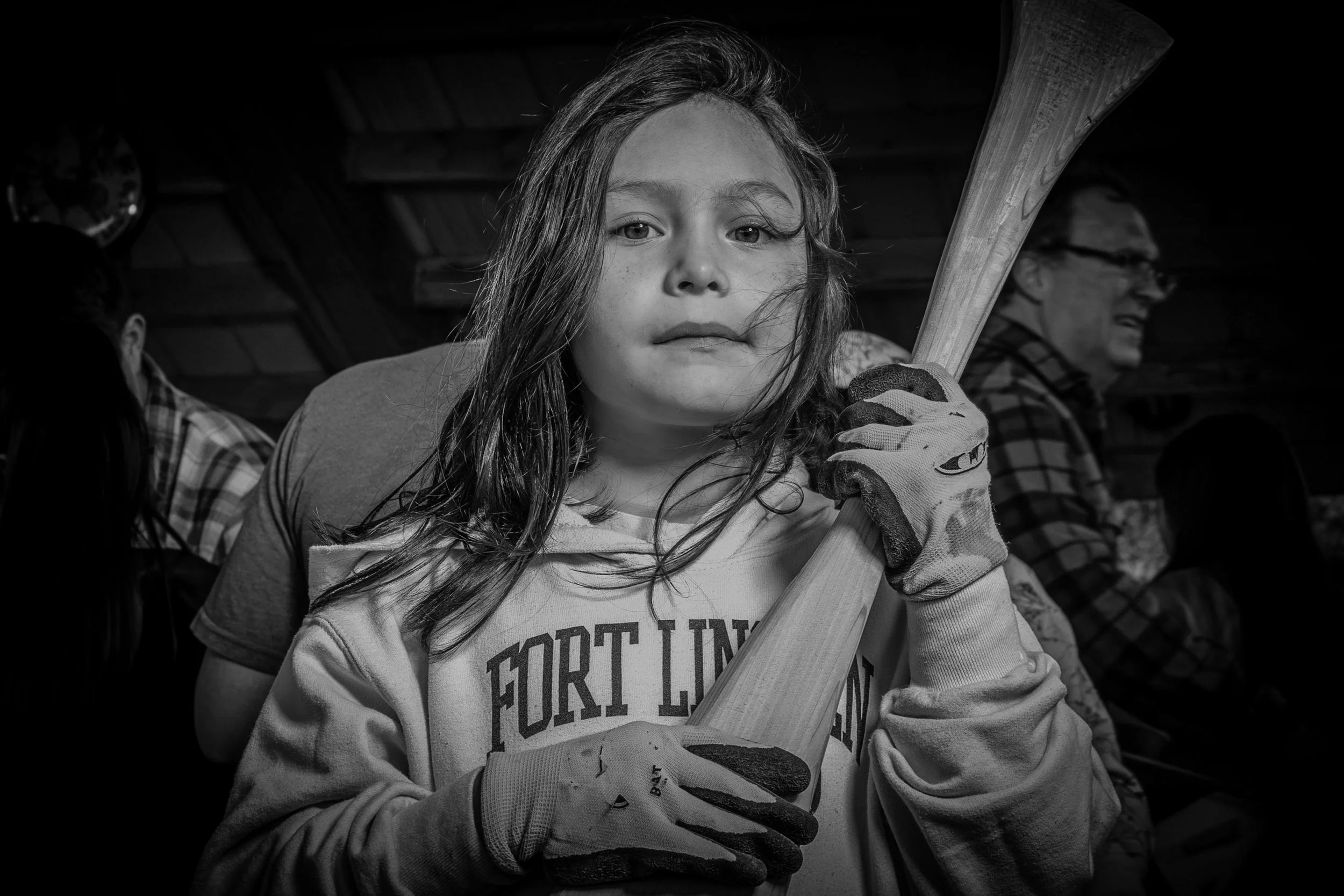 A young girl with long hair holding a baseball bat, wearing gloves, indoors. Two men are visible in the background, one wearing glasses.