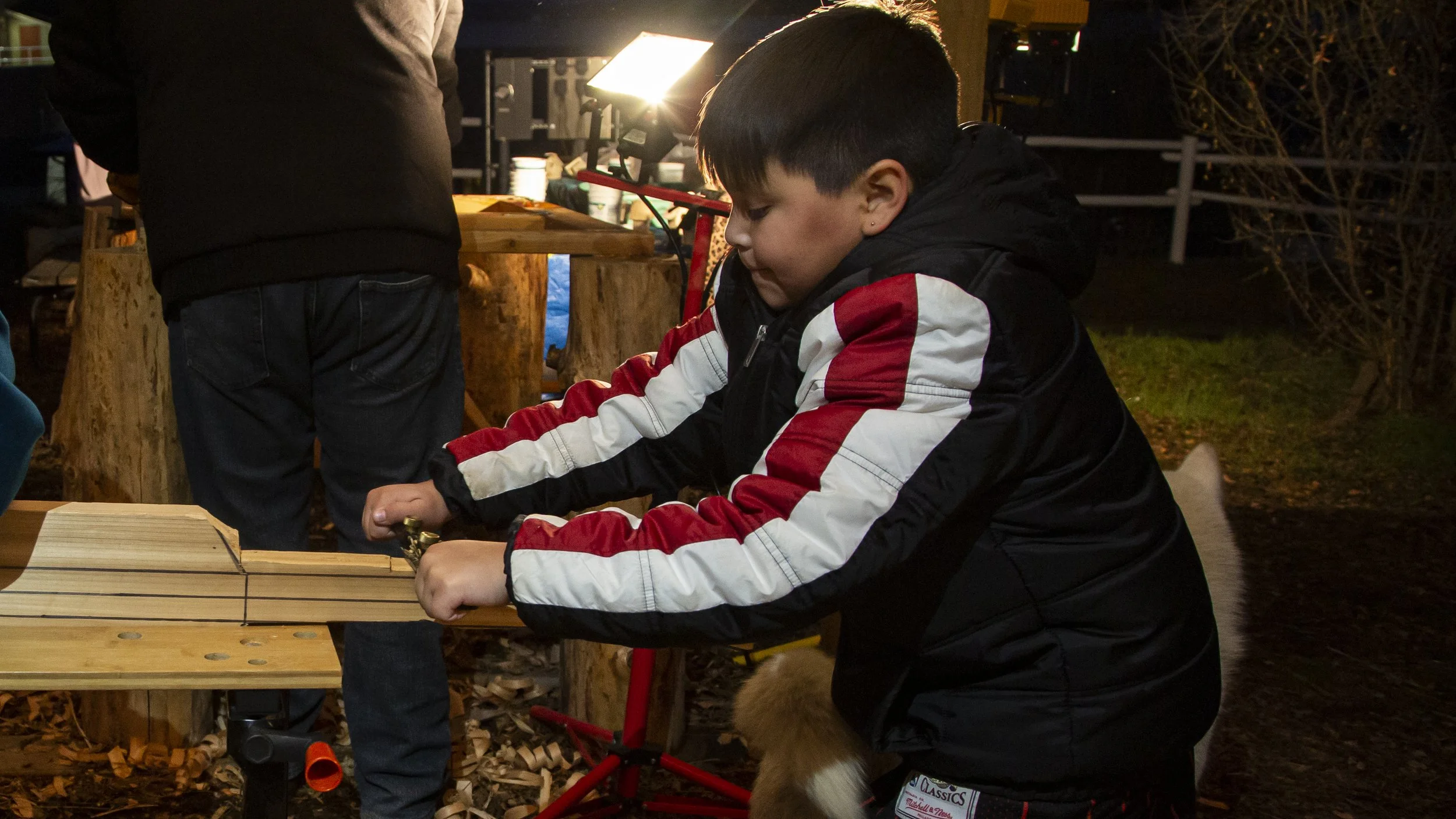 A young boy wearing a black jacket with red and white stripes, focused on working on a wooden project outdoors at night. The scene is illuminated by a bright light, with woodworking tools and wood shavings around.