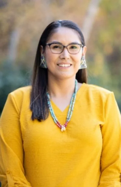 A woman with glasses and long dark hair wearing a yellow top and colorful jewelry, standing outdoors with autumn trees in the background.