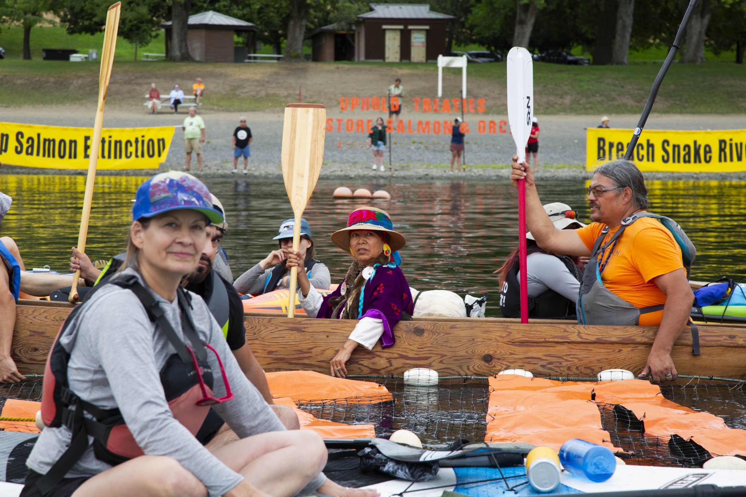 People in a boat on a river during daytime, with some holding paddles, surrounded by orange floating barriers, and a shoreline with signs and benches in the background.