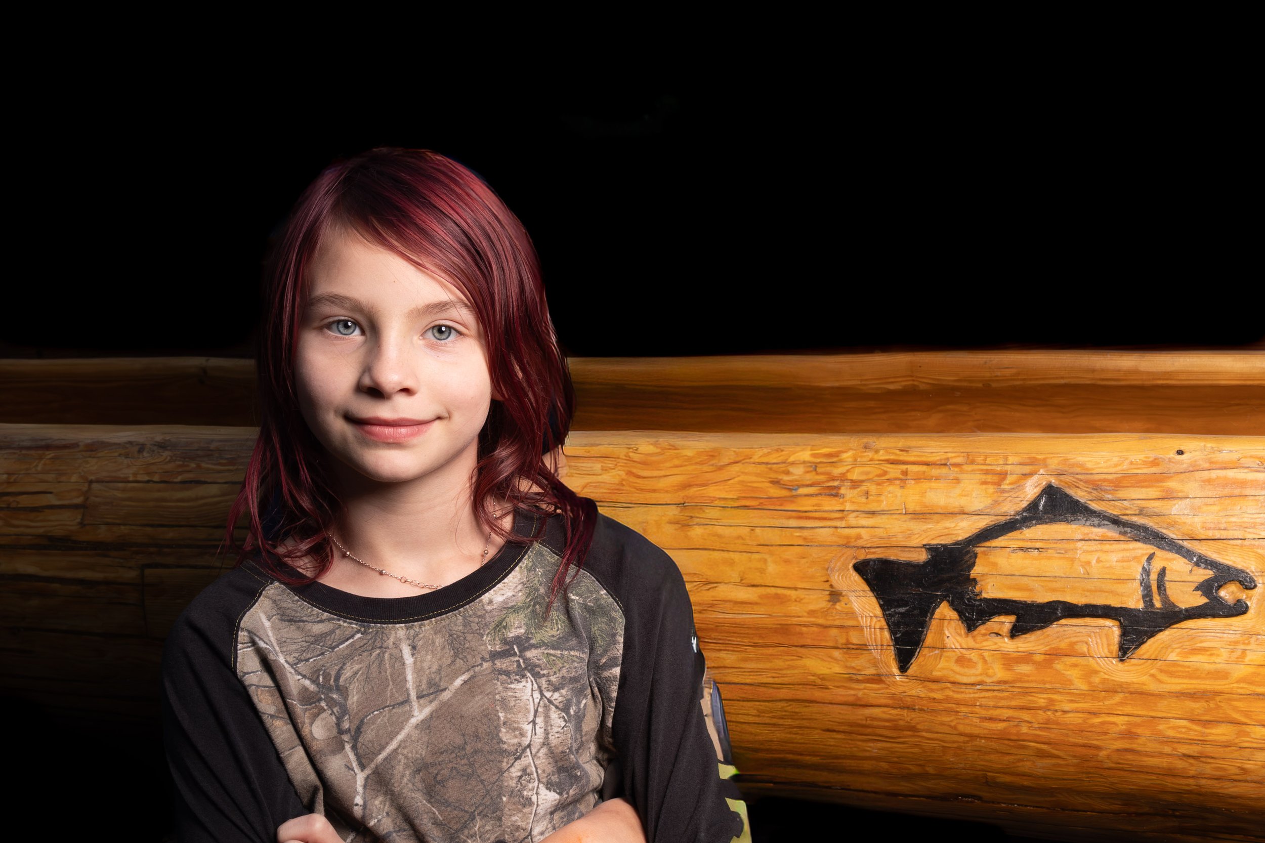 A young girl with red hair and blue eyes, wearing a camouflage-patterned shirt and a black jacket, stands in front of a wooden wall with black carvings.