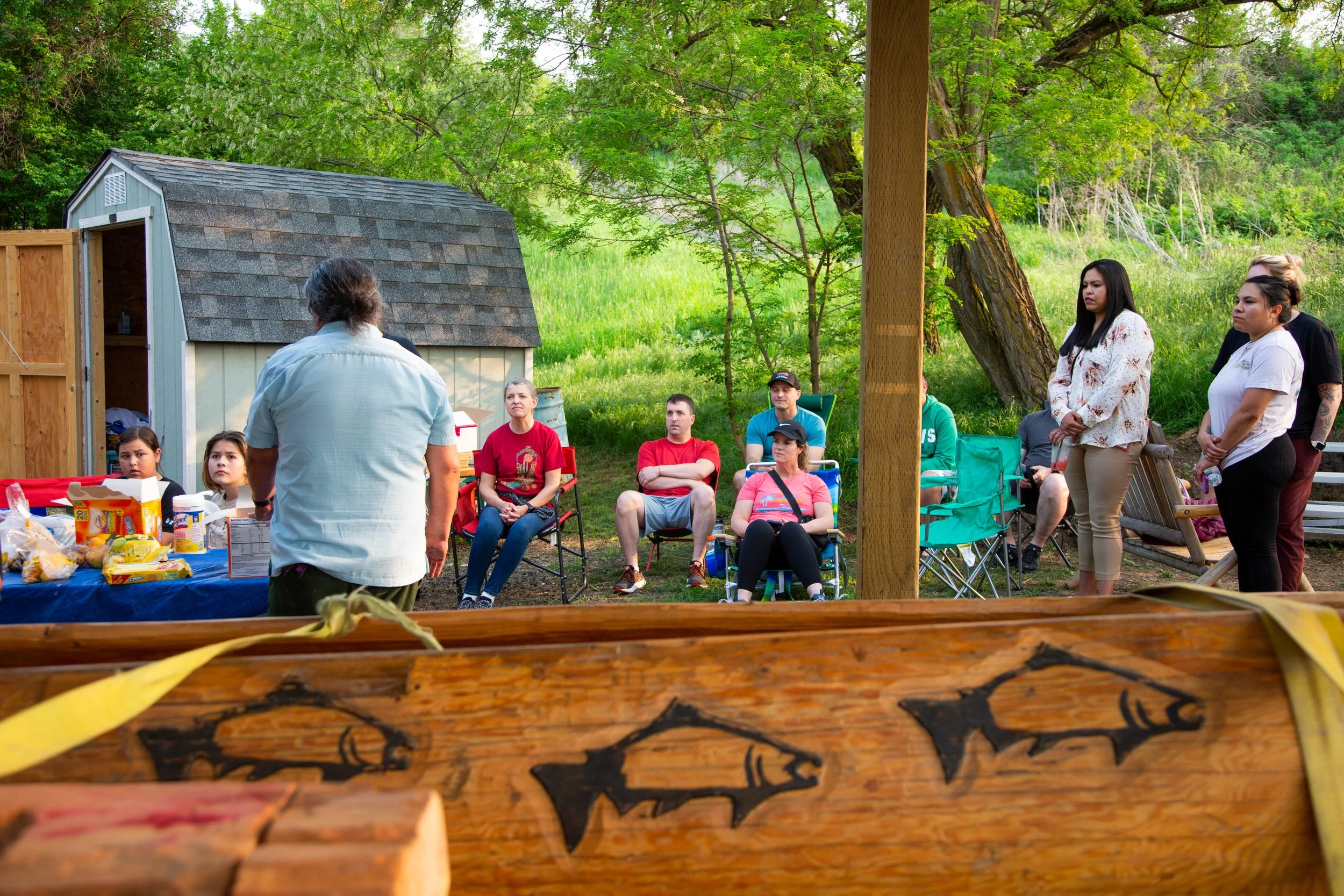 A group of people attending an outdoor gathering or meeting in a lush green park with trees and grass. They are sitting on chairs and standing around a table with food, listening to a person speaking.