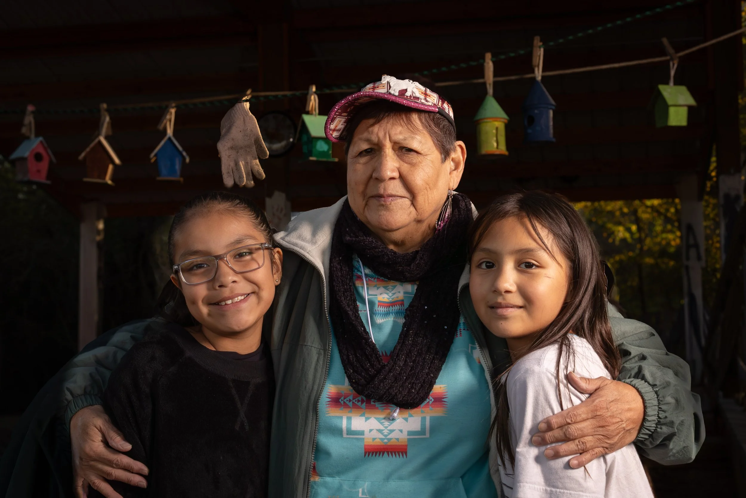 An older woman with short gray hair wearing a colorful hat and earrings, standing outdoors with childhood friends. Two young girls, one with glasses and the other with long brown hair, are embracing her and smiling. They are near a structure decorated with hanging birdhouses and gloves.
