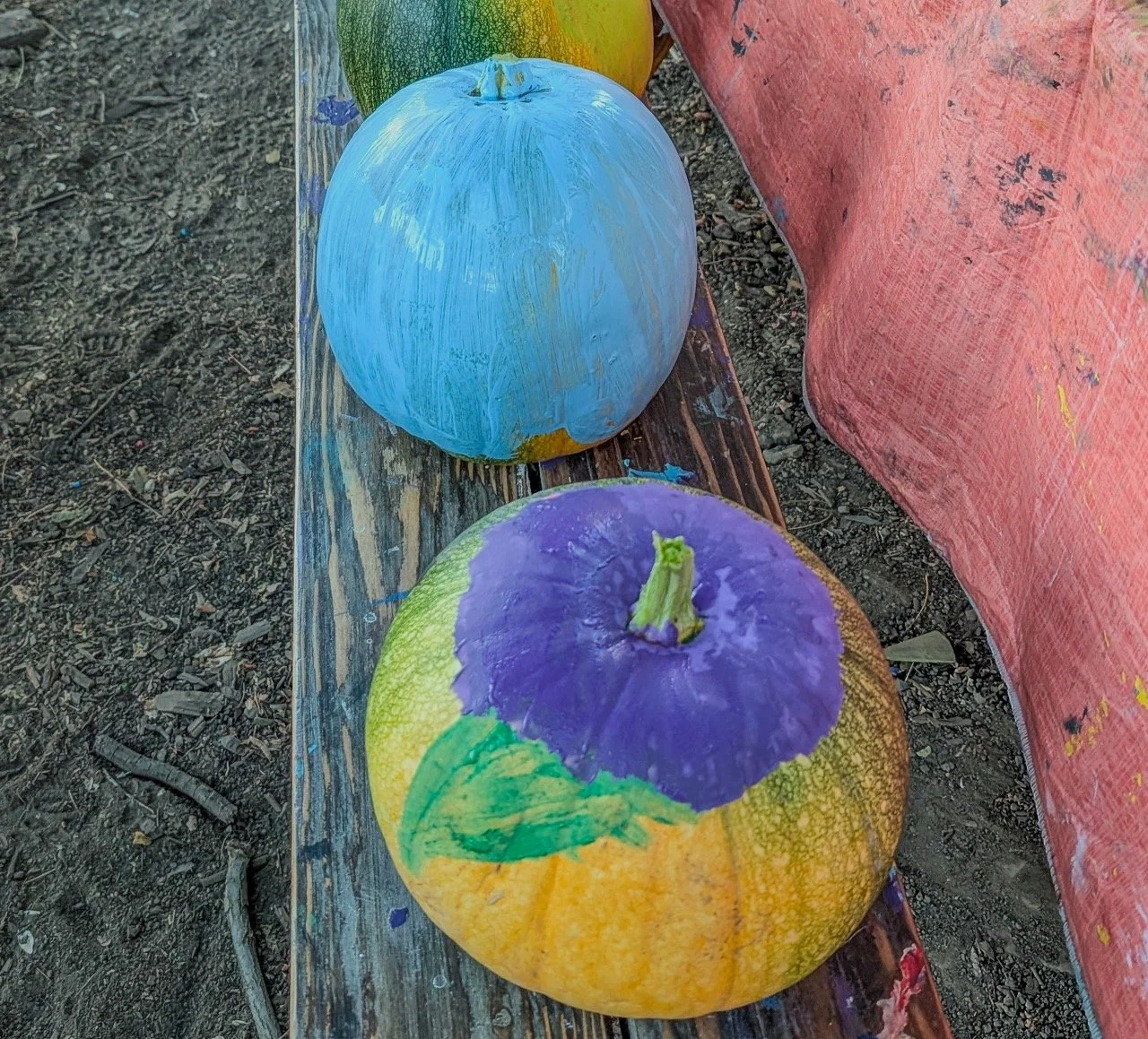 Two pumpkins painted with colorful designs, sitting on a wooden surface outdoors.