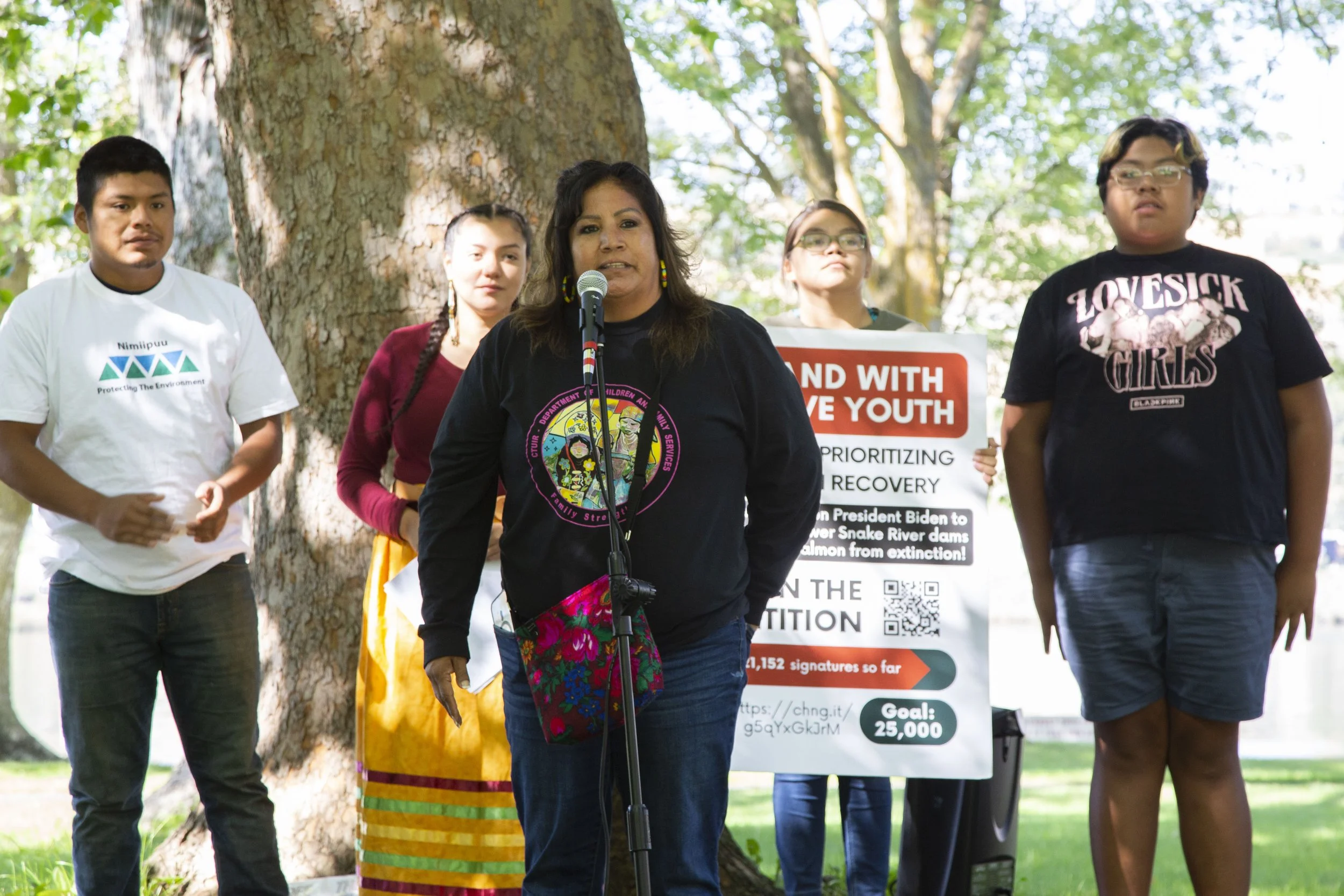 A group of people standing outdoors near a large tree during a protest or rally. One woman is speaking into a microphone, and others are holding signs supporting environmental and social causes.