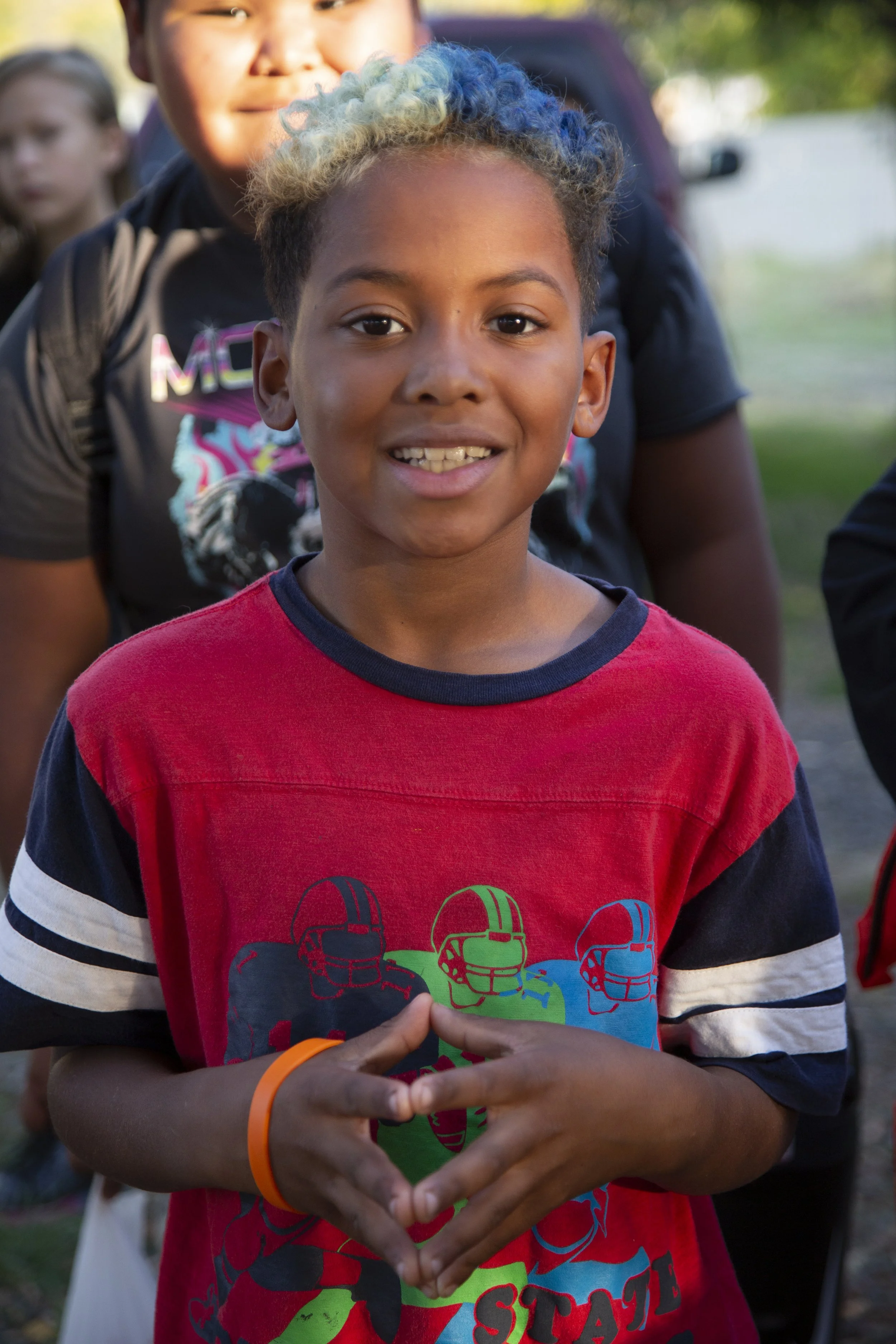 Close-up of a smiling young boy with curly, multi-colored hair, wearing a red T-shirt with graphic of football helmets and making a heart shape with his hands.