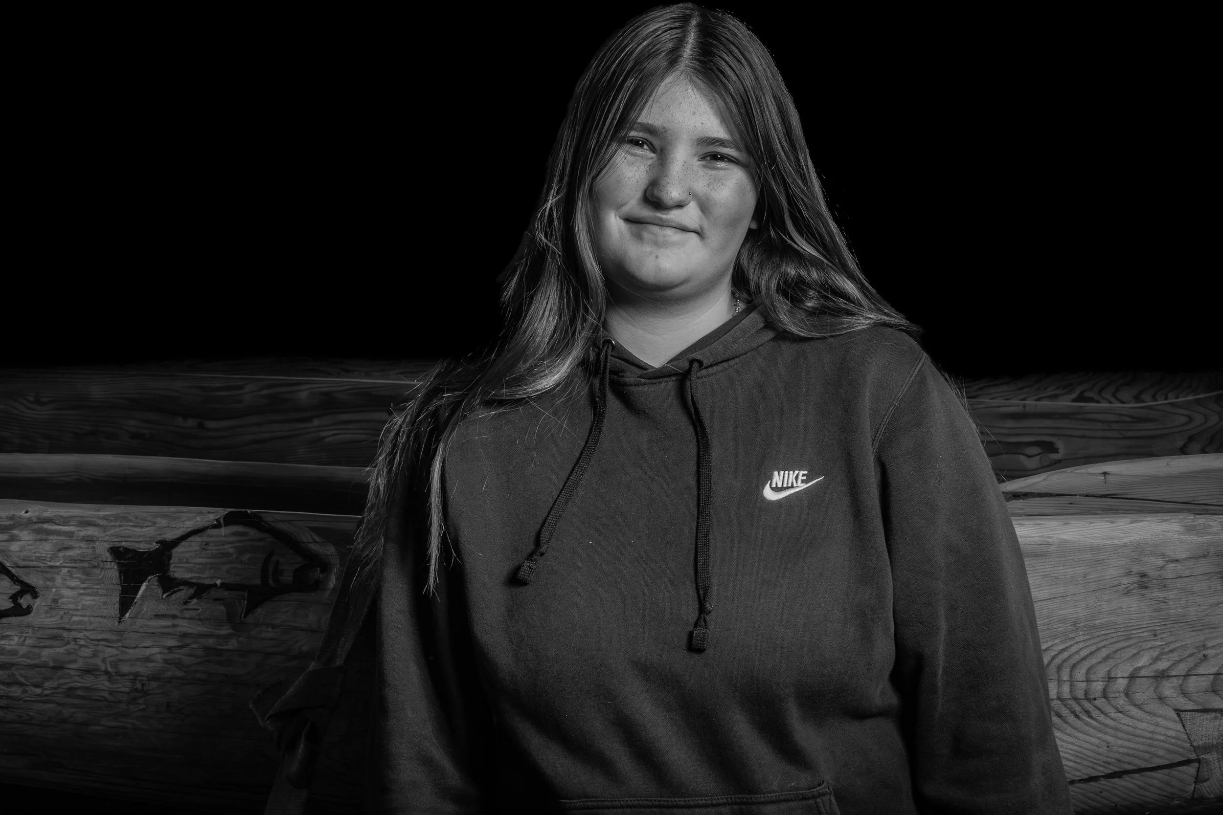 Black and white photograph of a young woman with long hair, wearing a hoodie with Nike logo, standing outdoors in front of a wooden fence.