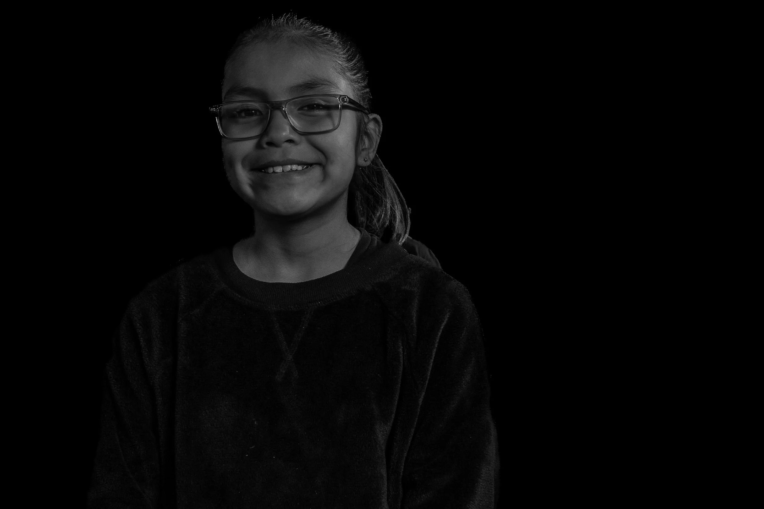A young girl with glasses smiling at the camera, wearing a dark sweater, against a black background.