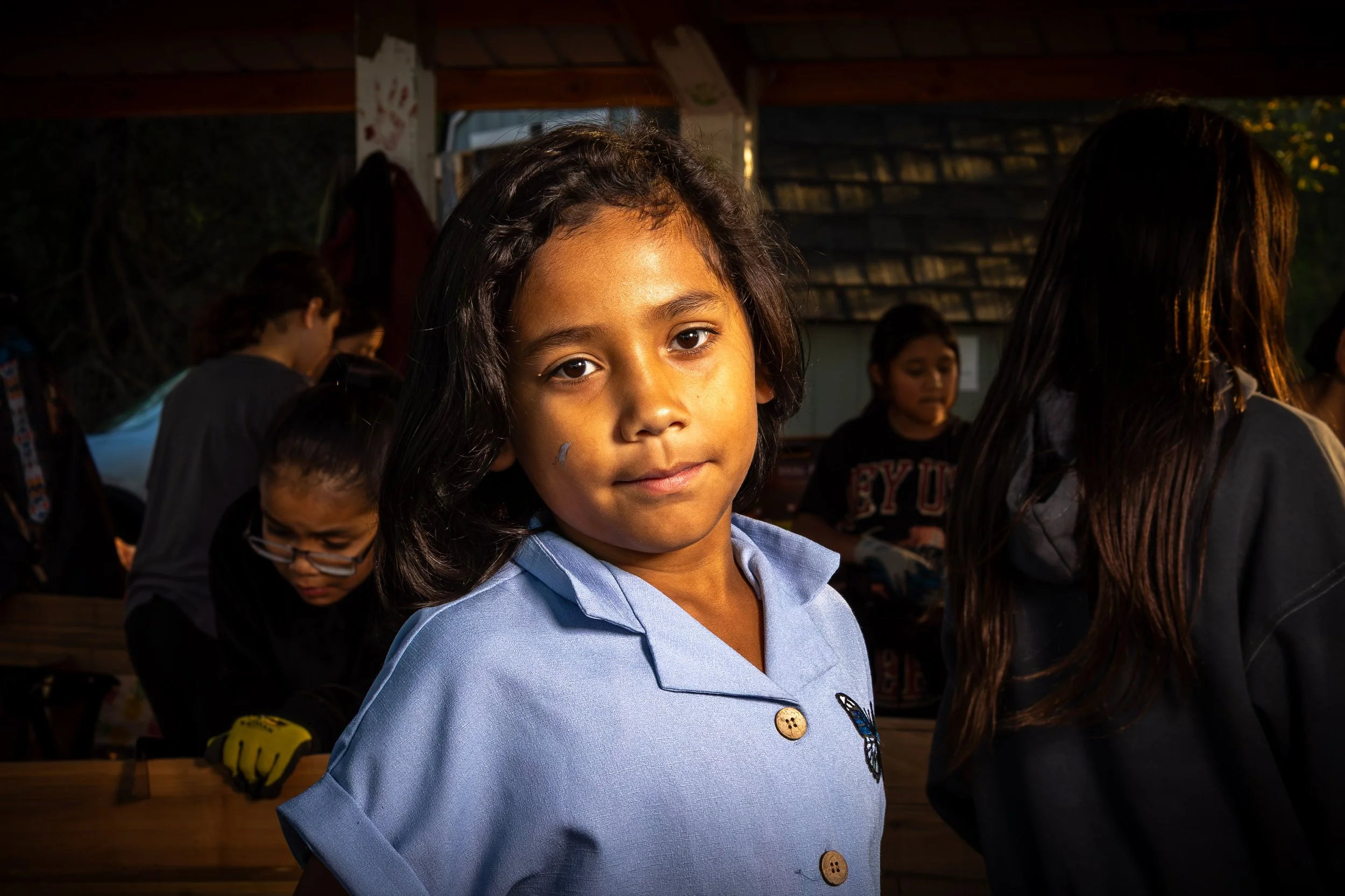 Young girl with dark hair wearing a light blue shirt, standing indoors with other children in the background.
