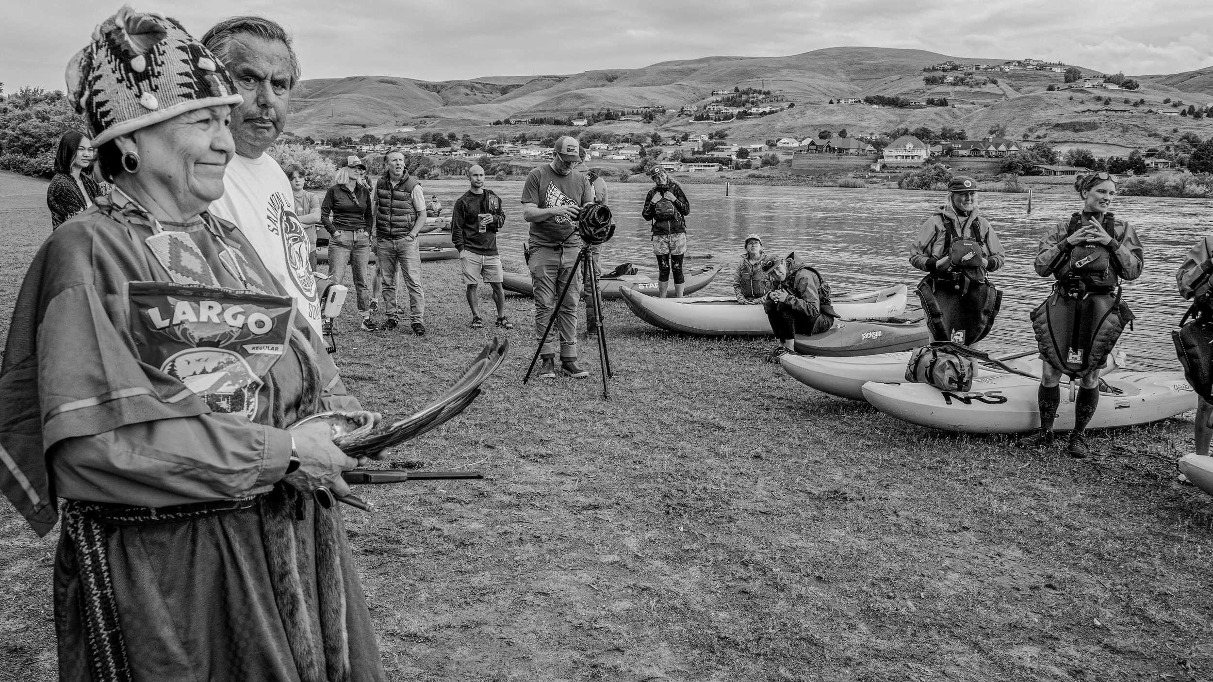 A group of people by a river, with kayaks on the shore and hills in the background. Some are standing, and others are preparing for kayaking.