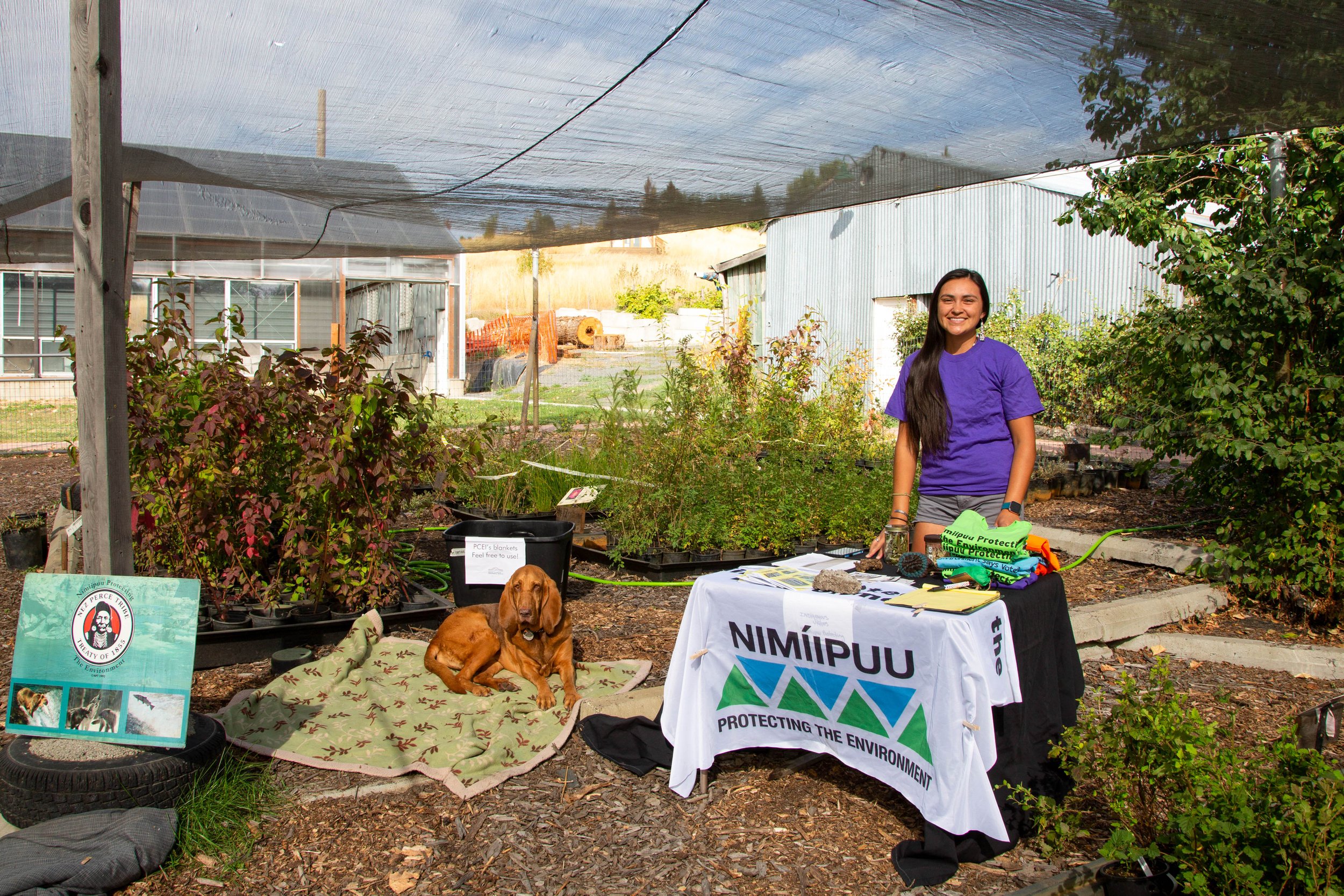 A smiling woman stands behind a table with environmental protection information and merchandise, under a black shade cloth in a garden. Two dogs are resting on a blanket in front of the table. Potted plants and signs are visible around the area.