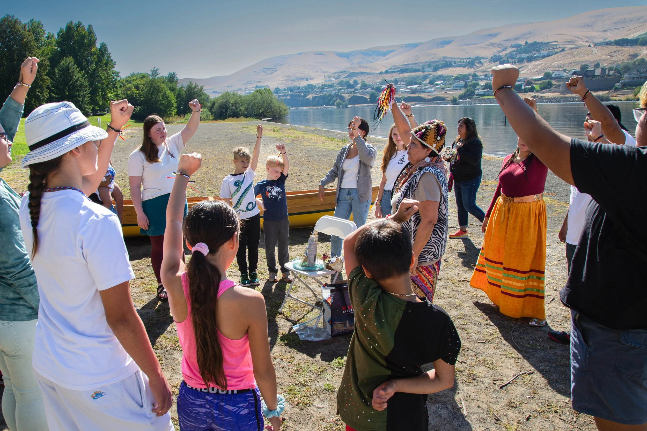 Group of people, including children and adults, gathered outdoors near a lake, engaging in a group activity with raised fists, on a sunny day with mountains and trees in the background.