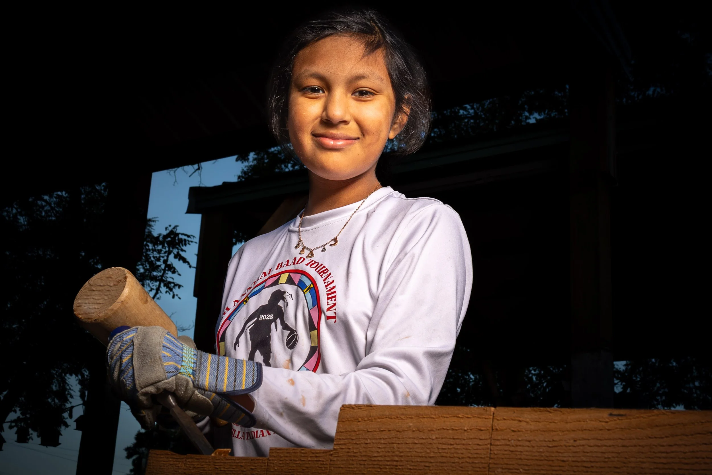 A young girl with short dark hair smiling at the camera, wearing a white T-shirt with a logo and necklace, gloves, and holding a hammer, working outdoors under a shaded structure with trees visible in the background.